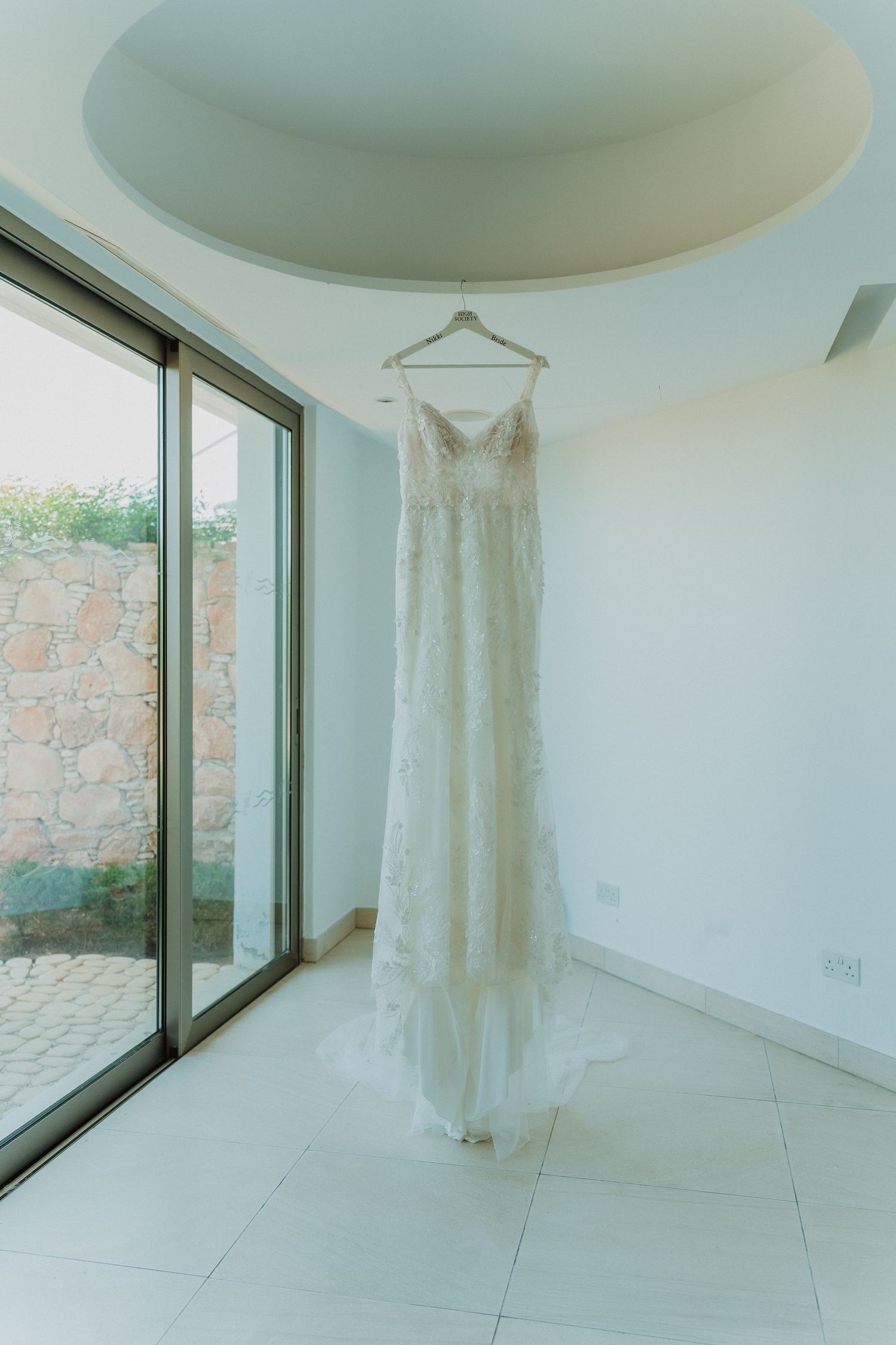 A white lace wedding dress hangs elegantly against a bright modern interior with large windows and white marble floors.