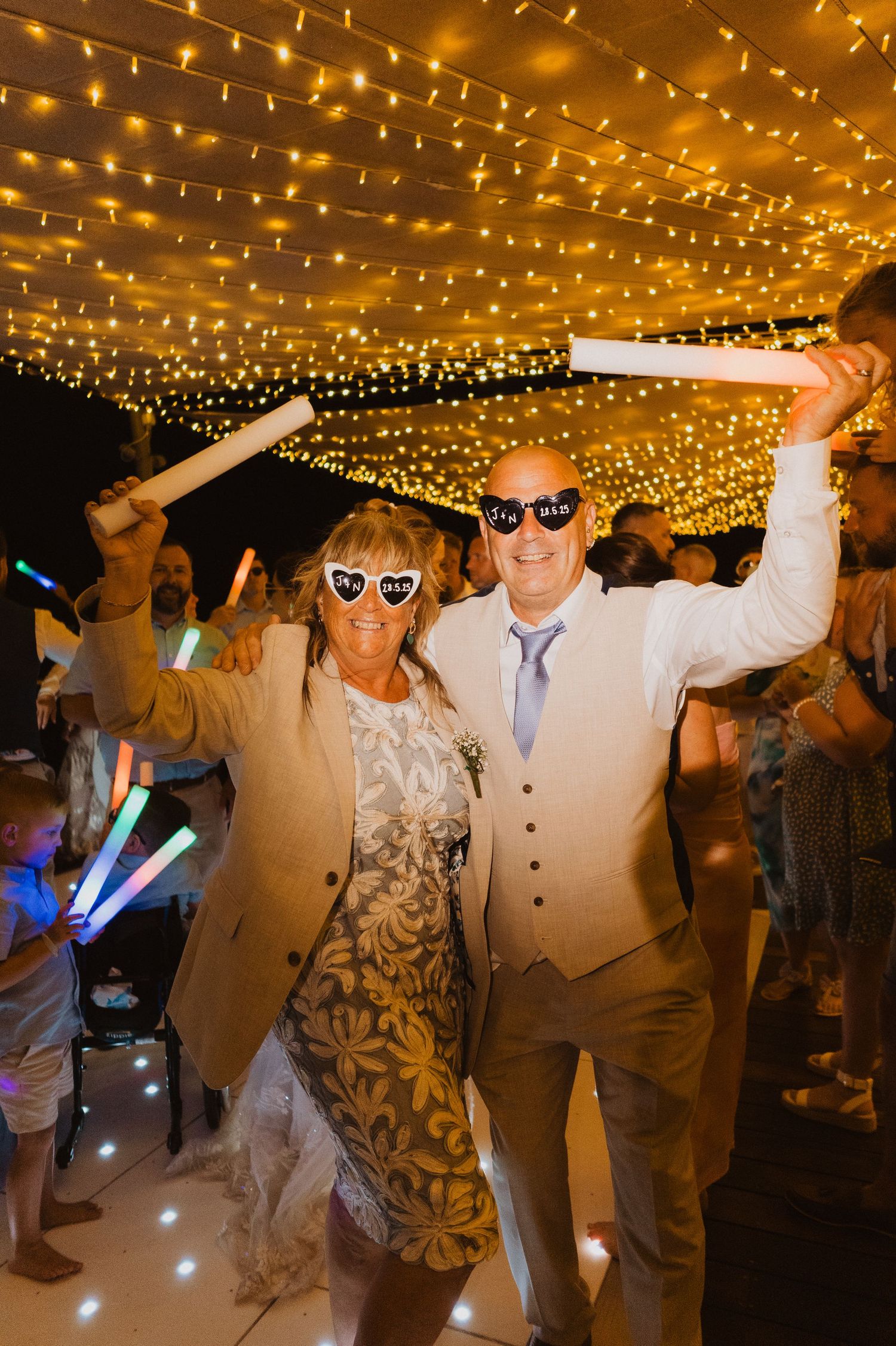 Wedding party dances under twinkling string lights at a reception with glowing LED accessories and festive atmosphere.