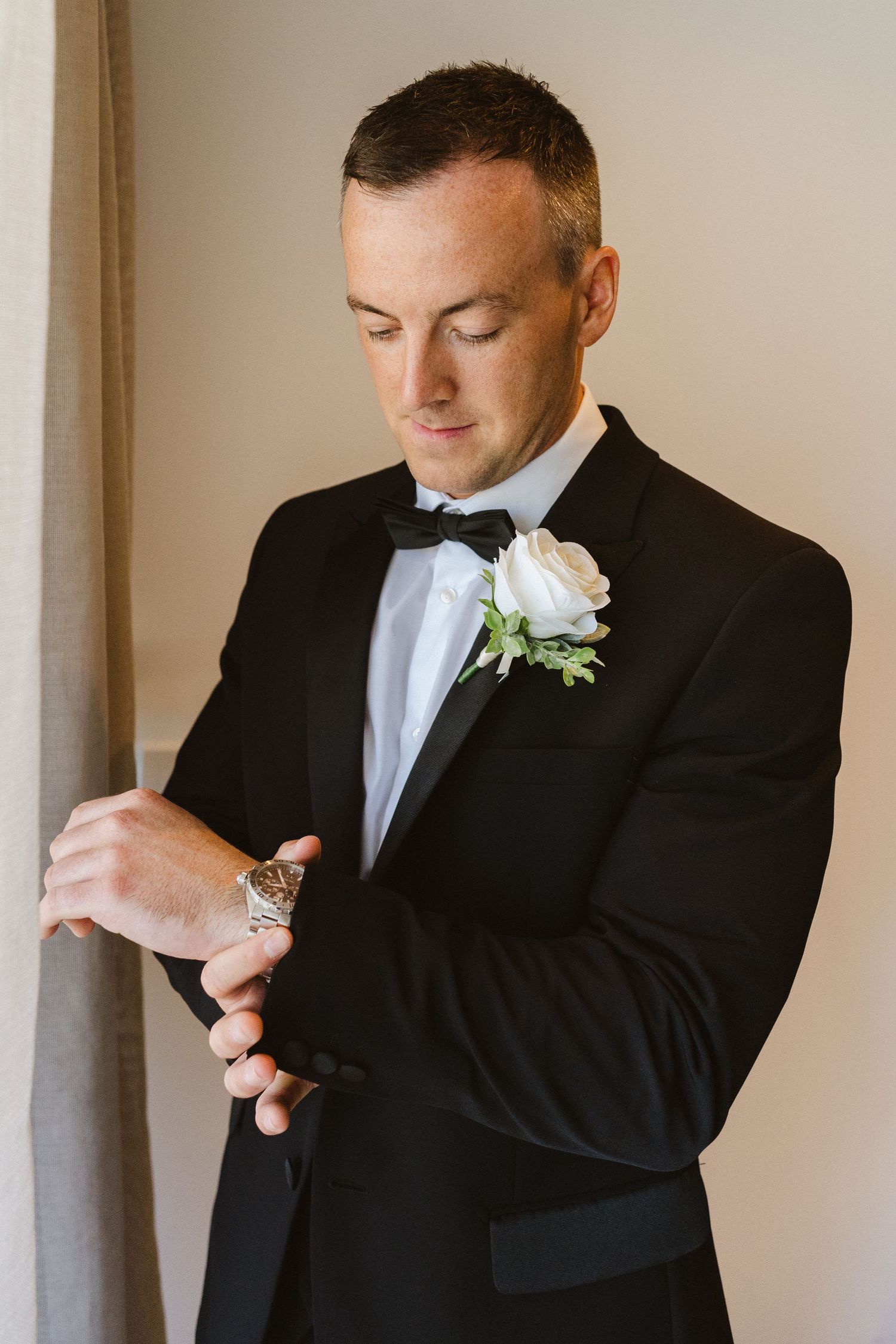 Groom in black tuxedo adjusts his tie while getting ready for wedding ceremony.