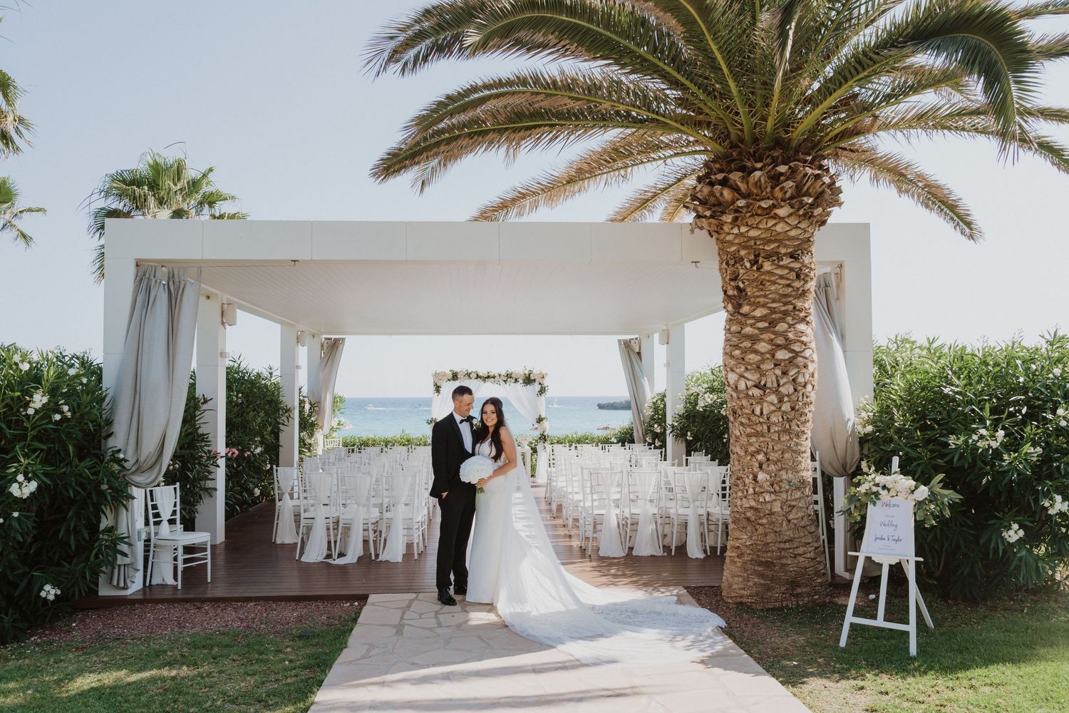 Elegant white wedding ceremony setup with palm trees and ocean views.