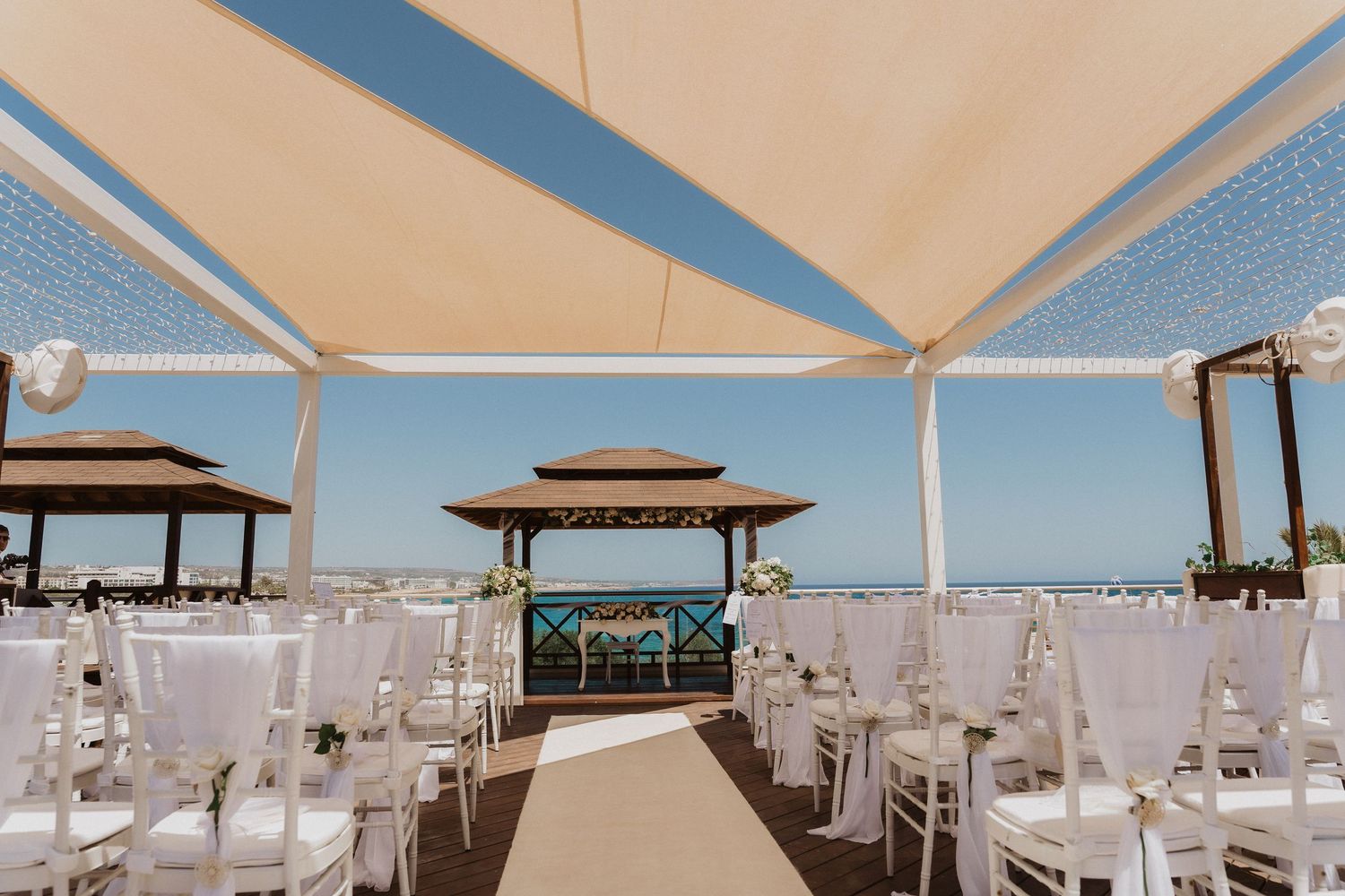 A panoramic view of an elegant beachfront wedding ceremony setup with white chairs and gazebos on a sunny day.