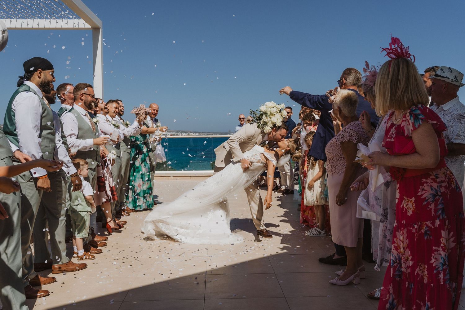 Wedding guests gathered on a sunny oceanfront terrace during a beach ceremony as a breeze catches the bride's veil.