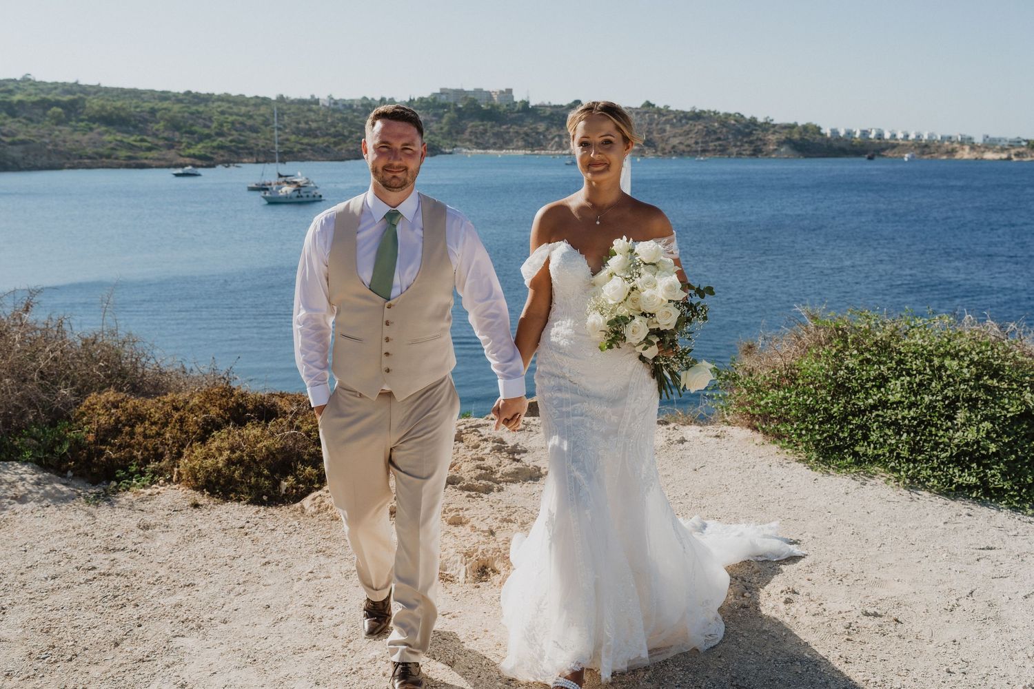 A couple walks hand in hand along a coastal path on their wedding day with a blue ocean and boats in the background.