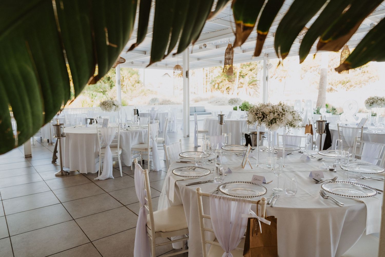 Elegant wedding reception setup with white tables and chairs under palm trees and string lights.