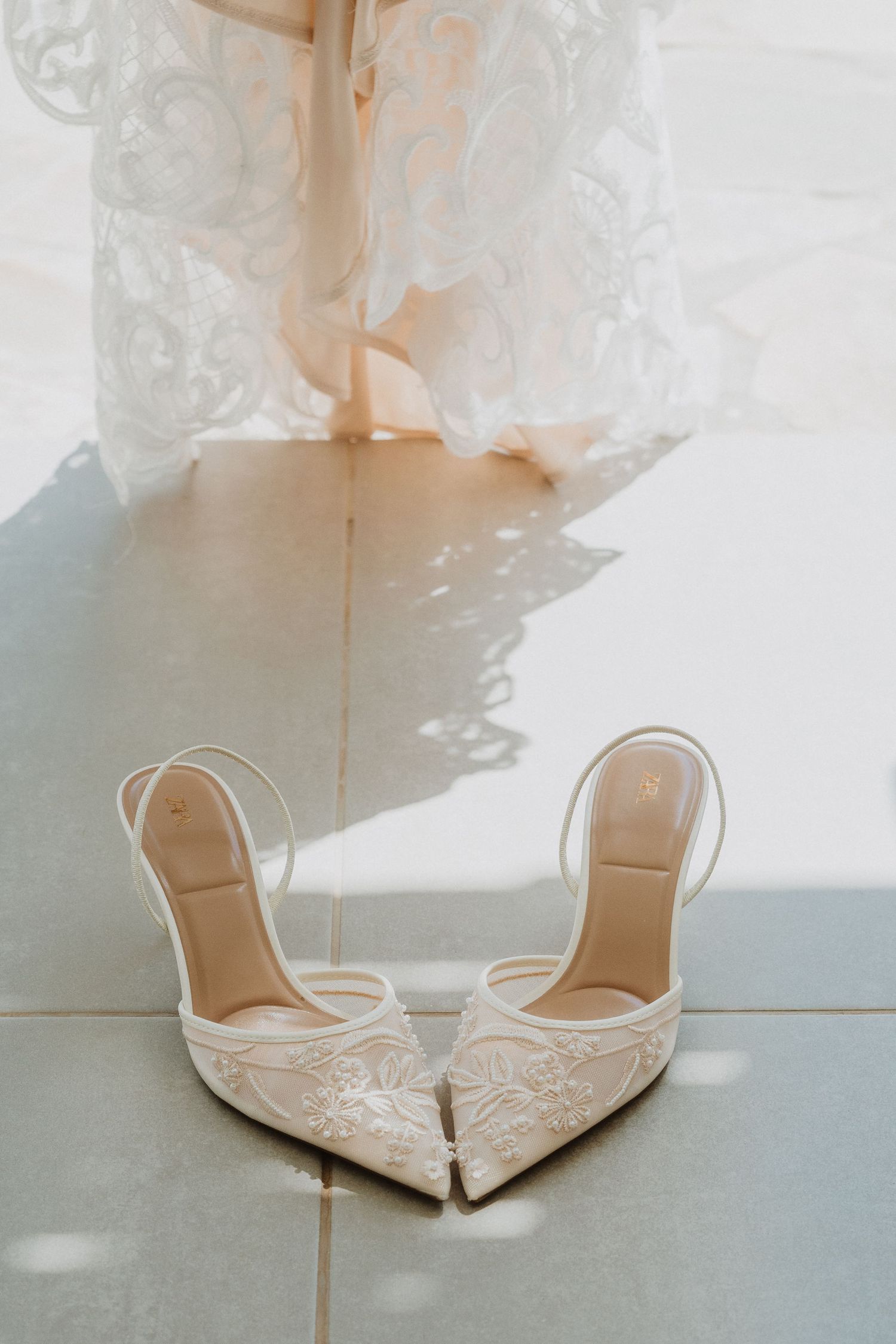 Elegant white lace bridal heels with pointed toes sit on a tiled floor beneath flowing fabric in soft natural light.