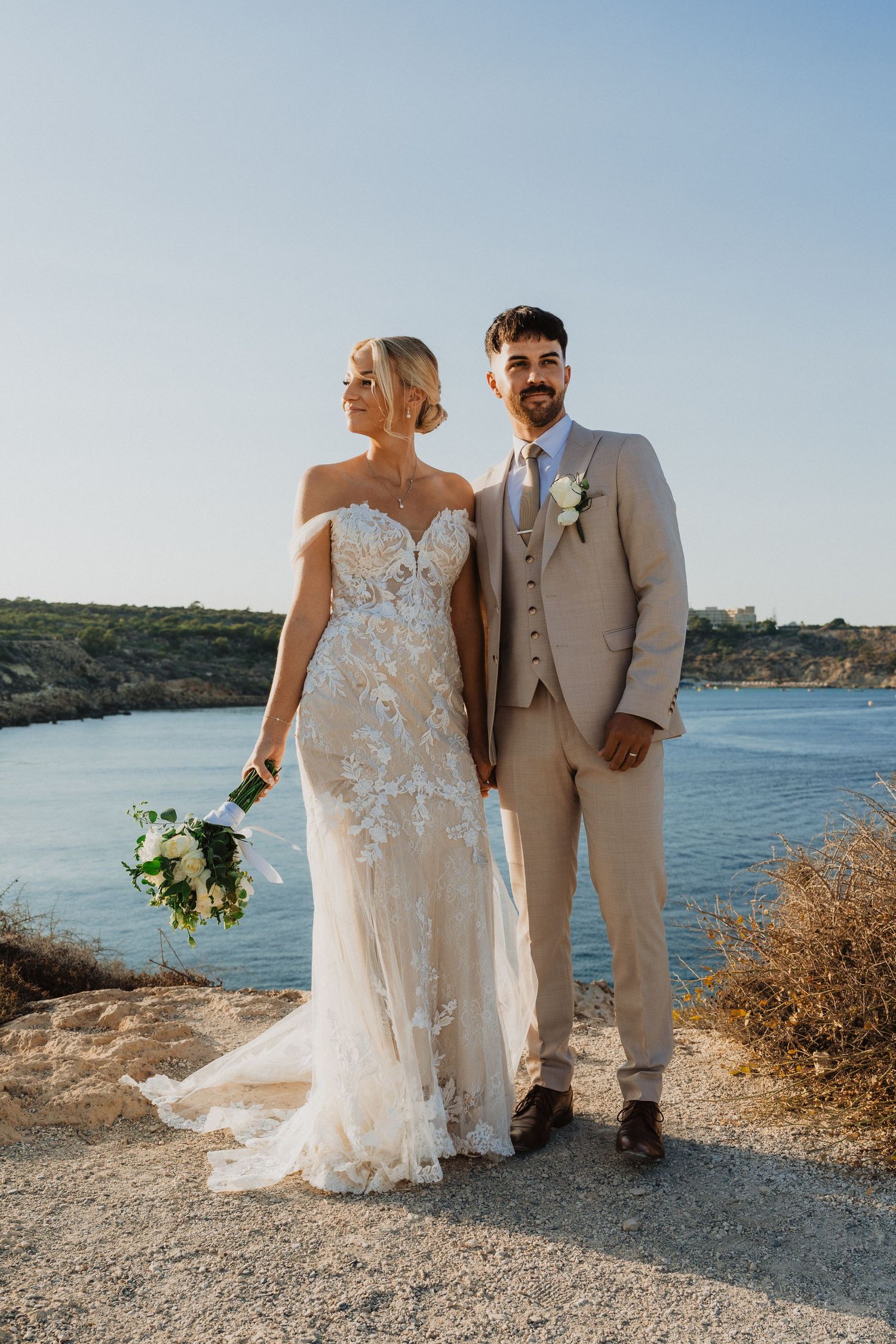 Wedding couple stands together on a scenic cliff path with water and coastline views in the background.