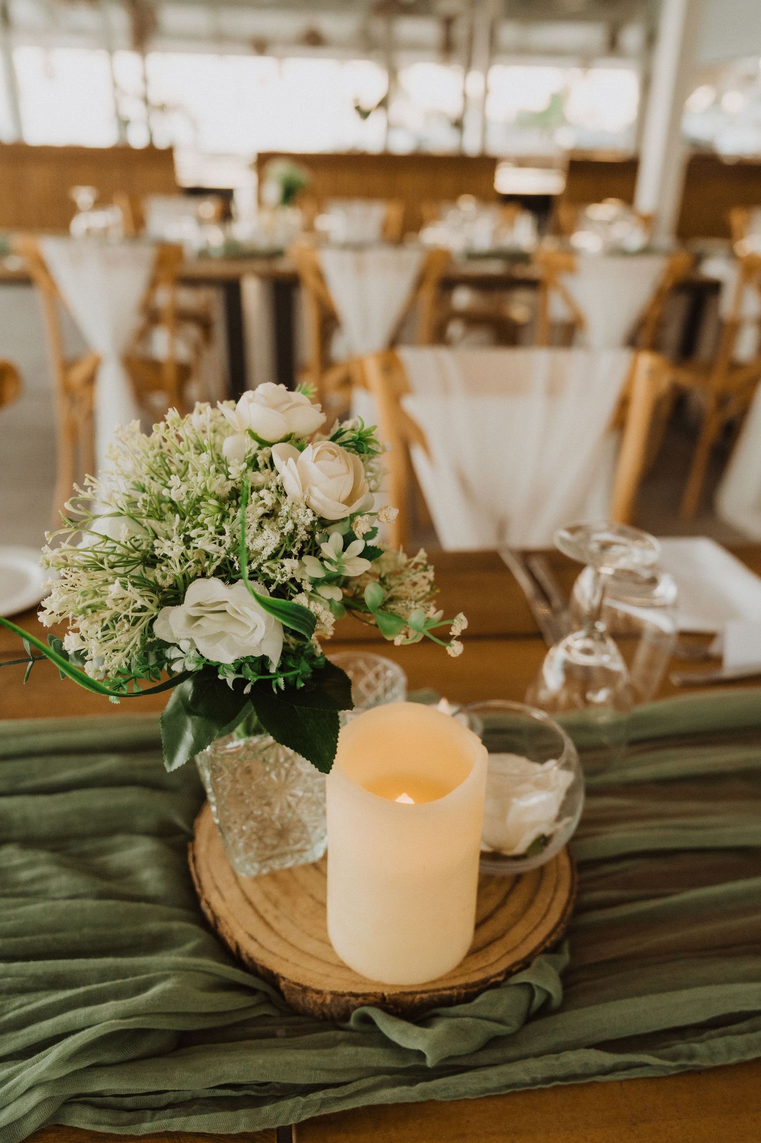 Rustic wedding centerpiece featuring white flowers and candles on wooden slice with green table runner.