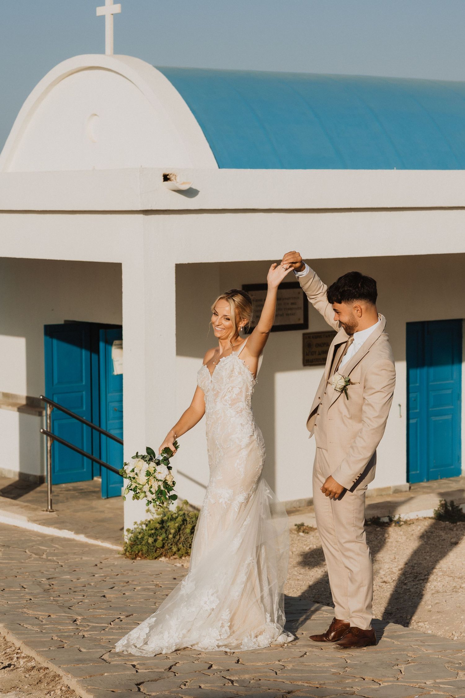 Wedding couple dances joyfully outside a white Mediterranean chapel with blue accents.