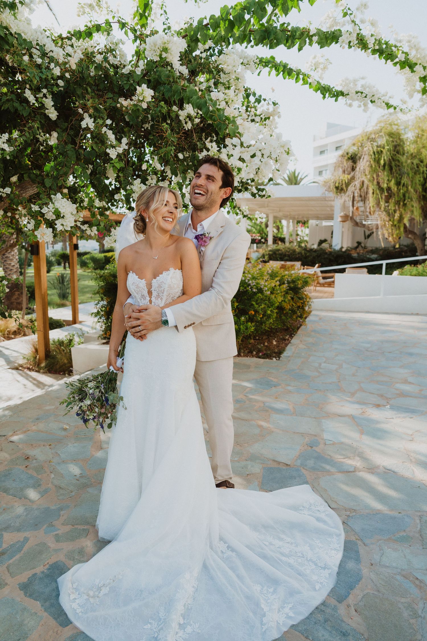 Bride in lace gown and groom in light suit embrace on stone patio under greenery.
