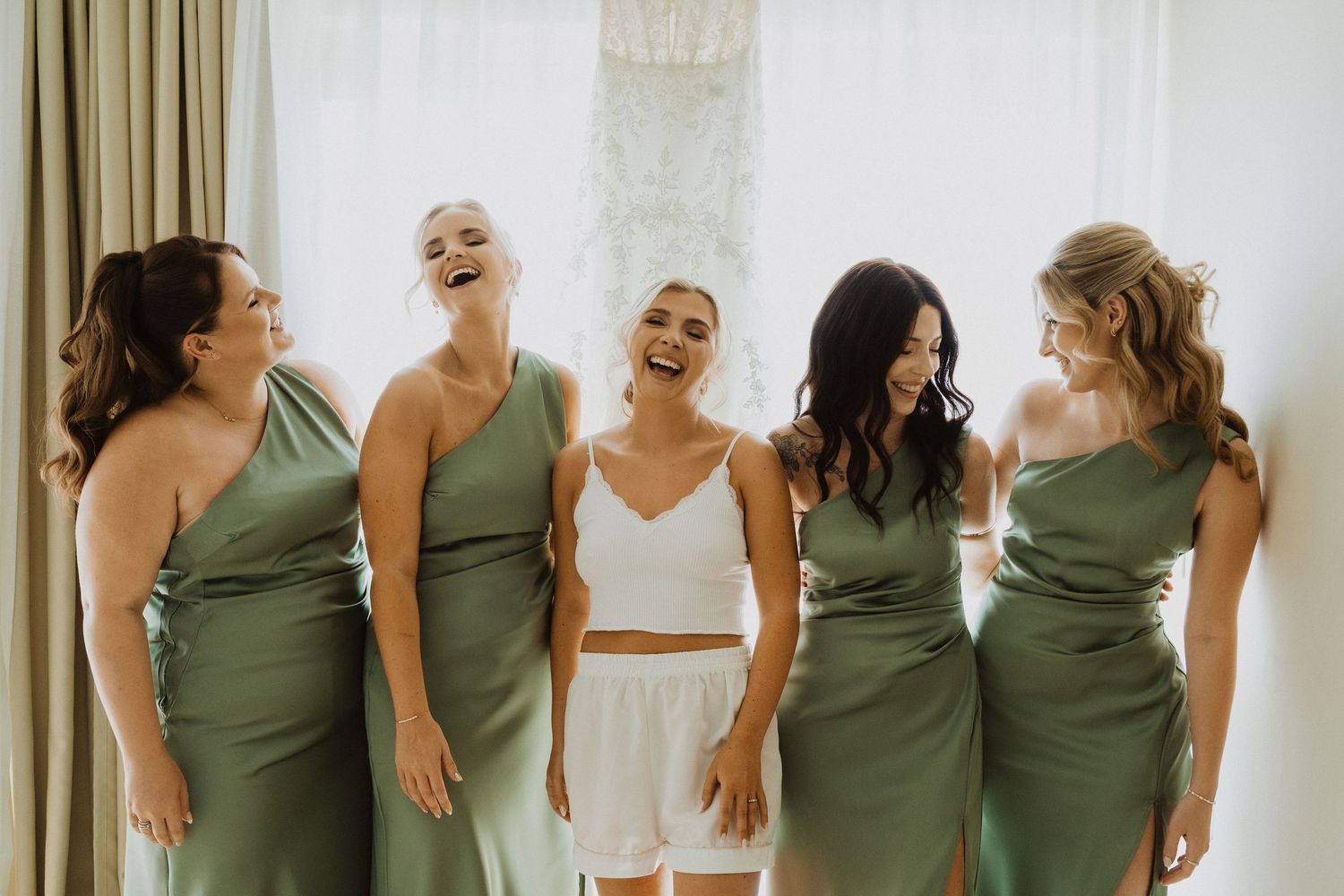 Wedding party in sage green dresses share a candid laughing moment together.
