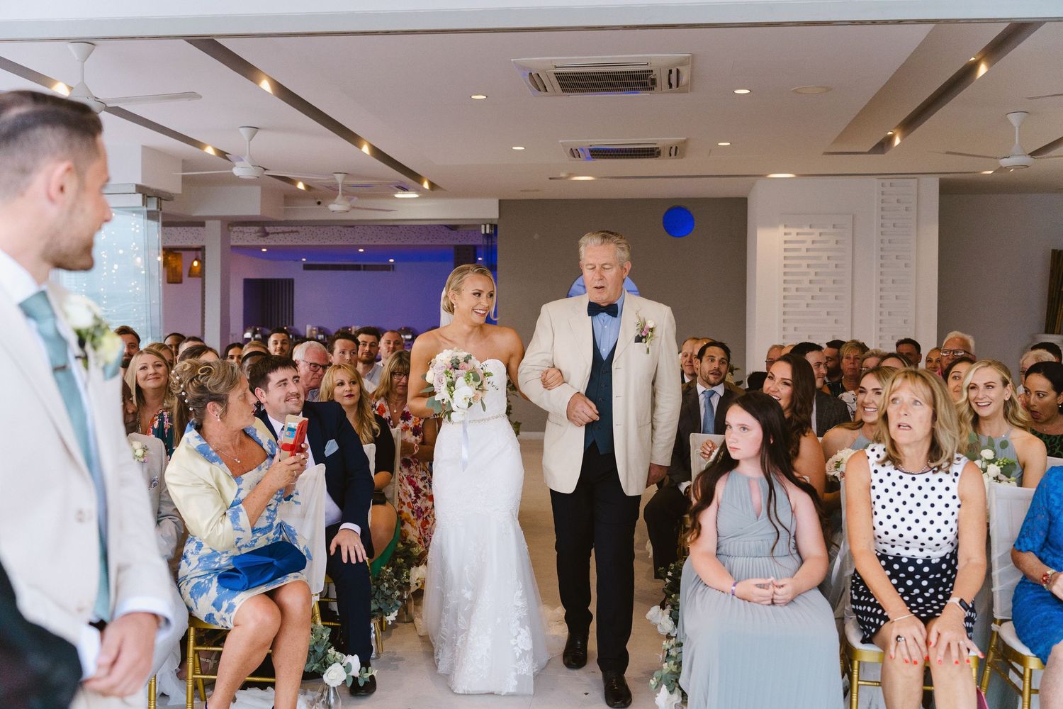 Indoor wedding ceremony with guests seated on white chairs and purple ambient lighting.