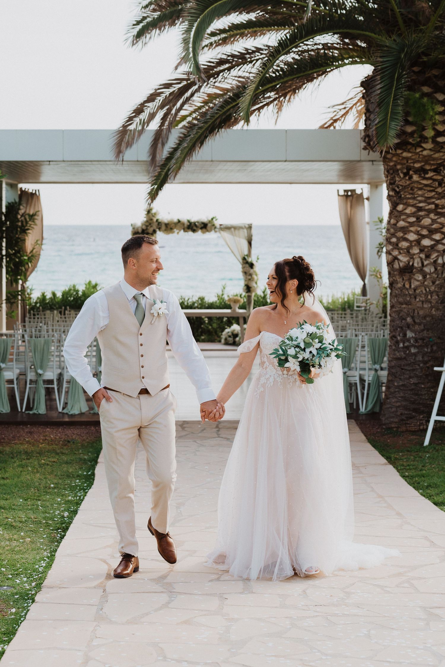 A couple holds hands during their beach wedding ceremony under a palm tree and floral arch by the ocean.