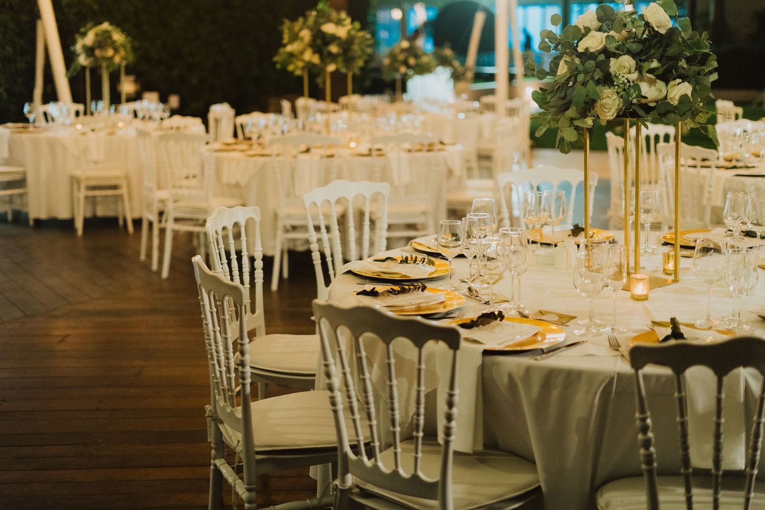 Elegant wedding reception tables set with white linens, gold accents and floral centerpieces in a banquet hall.