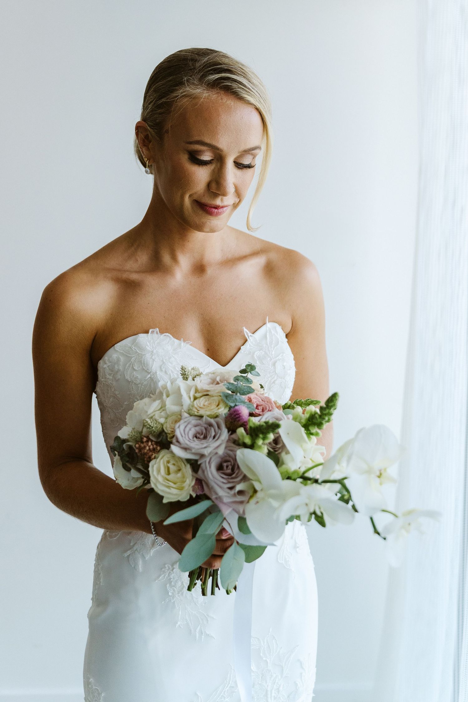 Bride in white lace dress gazes down at delicate pastel bouquet.