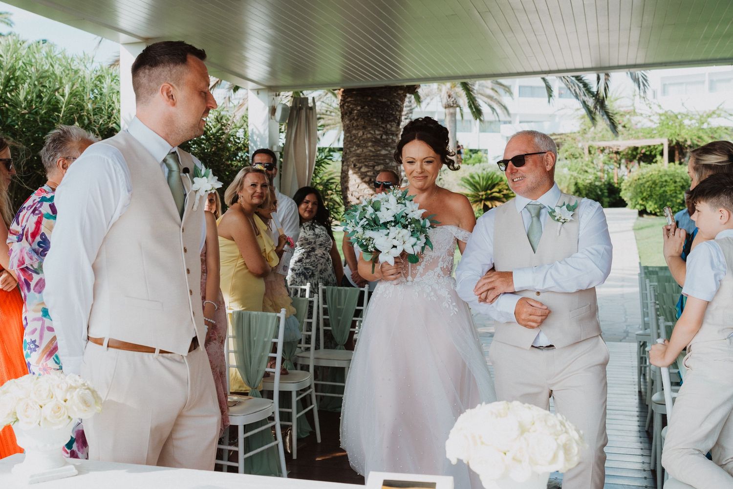 Wedding procession takes place on a covered porch decorated with white chairs and tropical greenery.