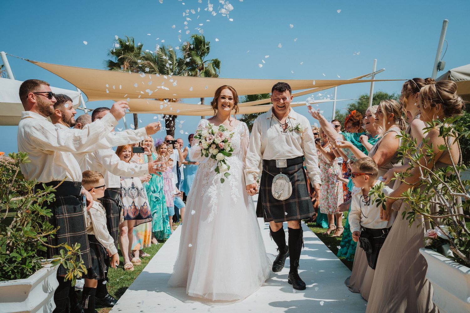 Joyful outdoor wedding ceremony with guests throwing confetti at couple under beige sail shades.