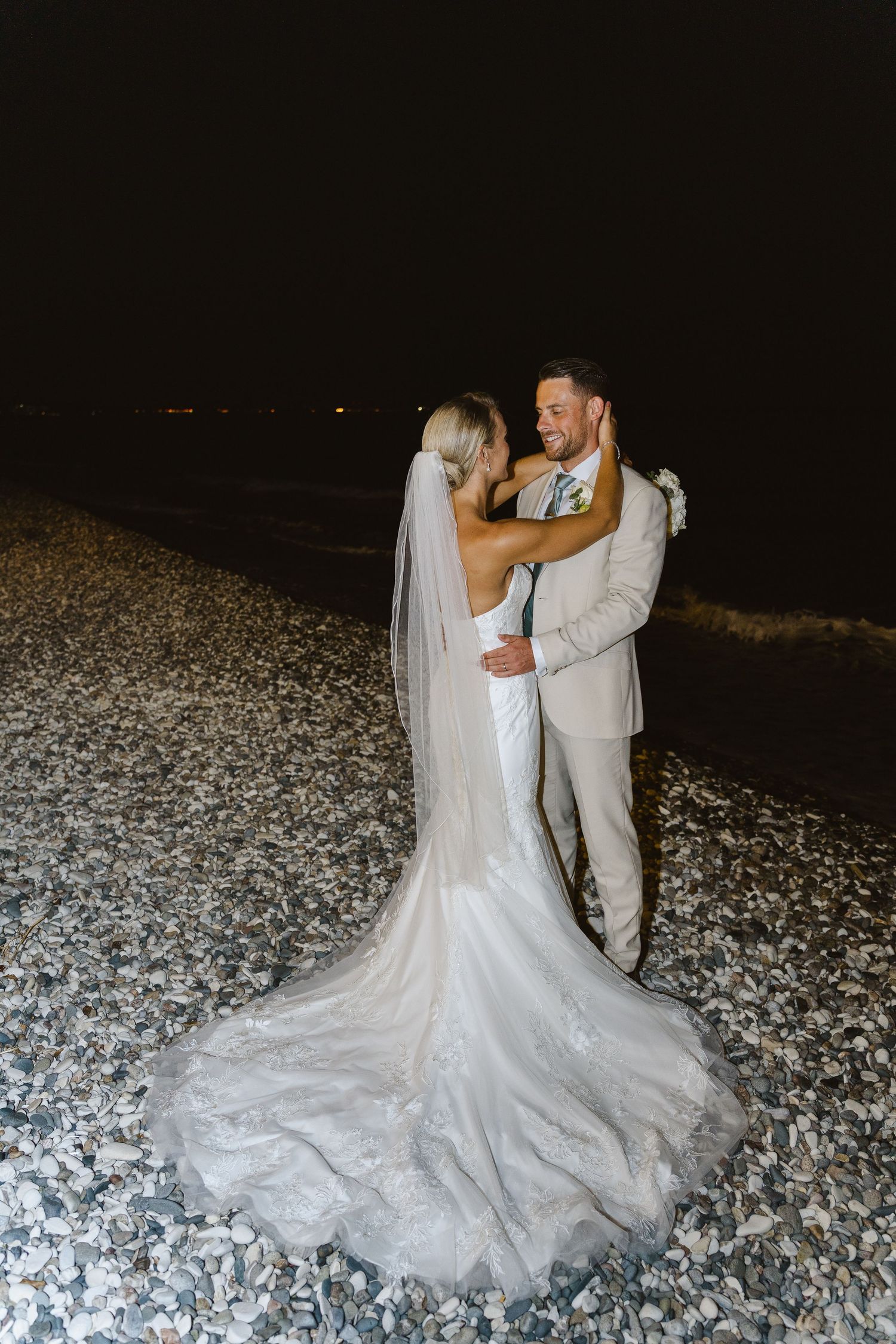Elegant white wedding dress with dramatic train spreads across rocky beach at night.