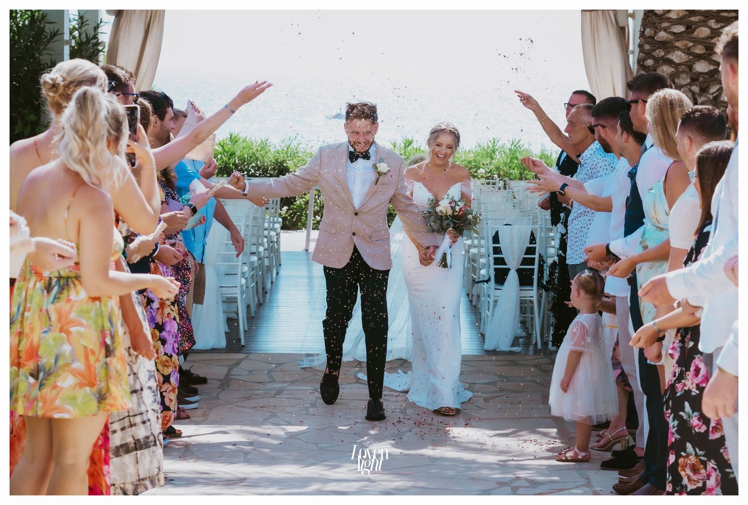 Wedding guests line both sides of an outdoor aisle as newlyweds joyfully walk between them.