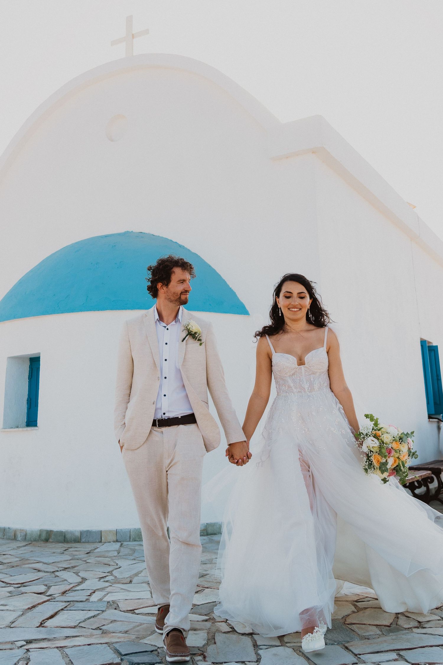 Joyful couple walking hand in hand by traditional Greek church architecture.