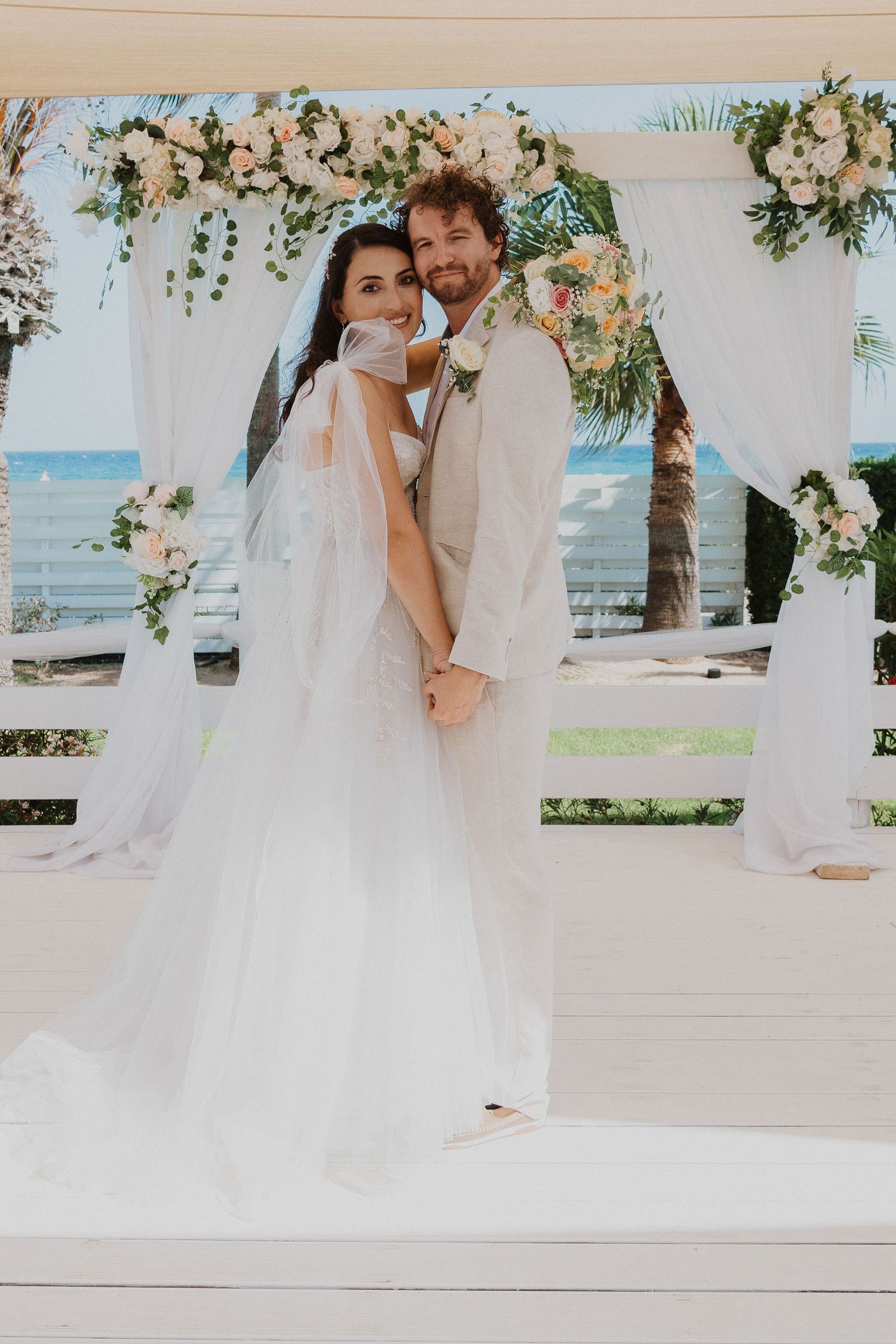 Beach wedding ceremony under a floral arch with white draping and ocean view in the background.