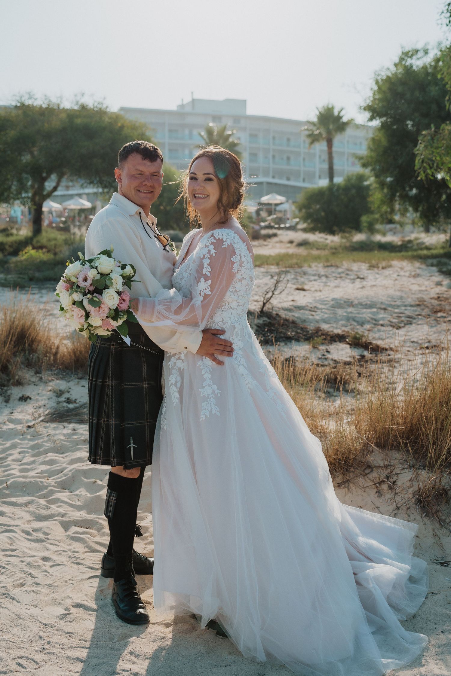 A wedding couple poses on a sandy beach with tropical palm trees and buildings in the background.