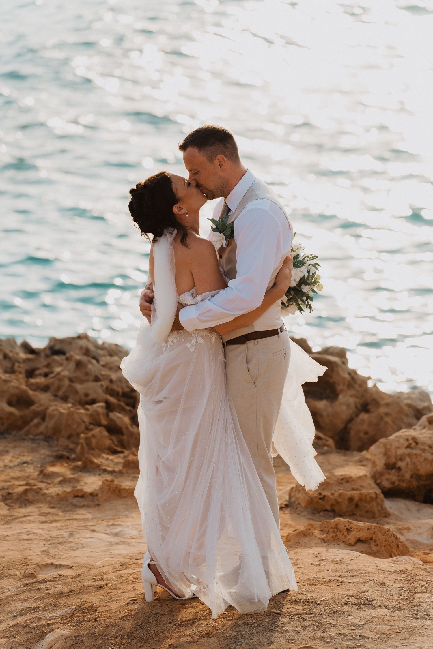 A romantic wedding couple embraces on rocky cliffs overlooking turquoise ocean waters at sunset.