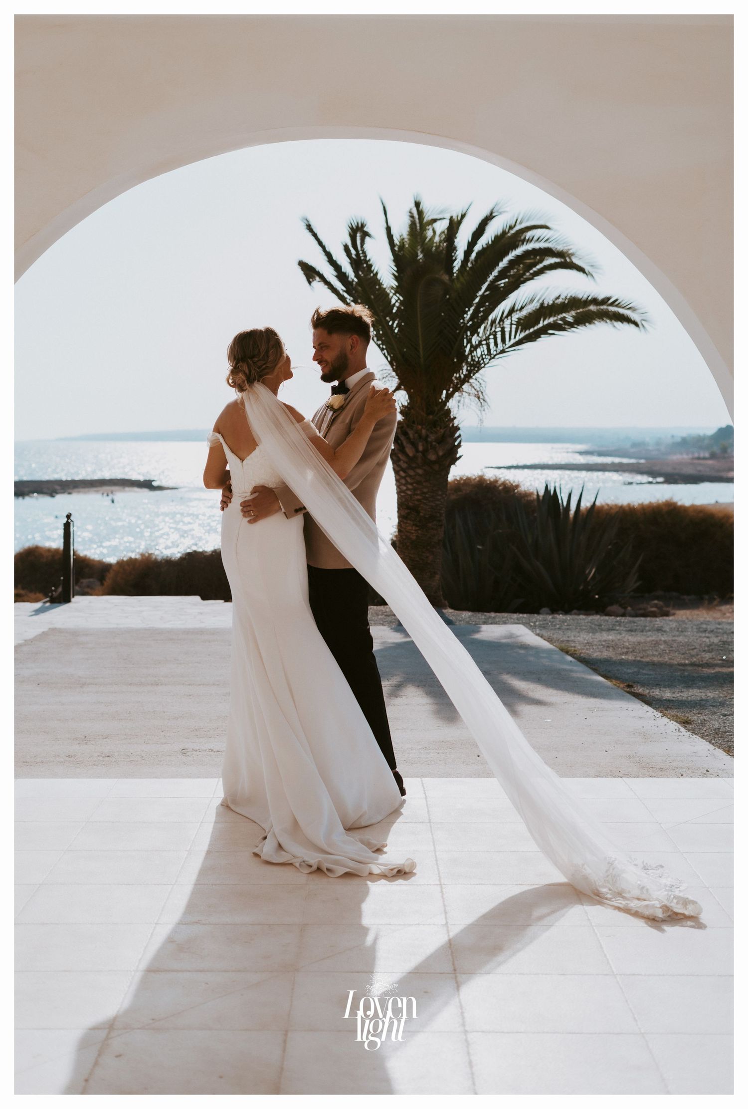 Wedding couple embraces under arched doorway with palm tree and ocean view.