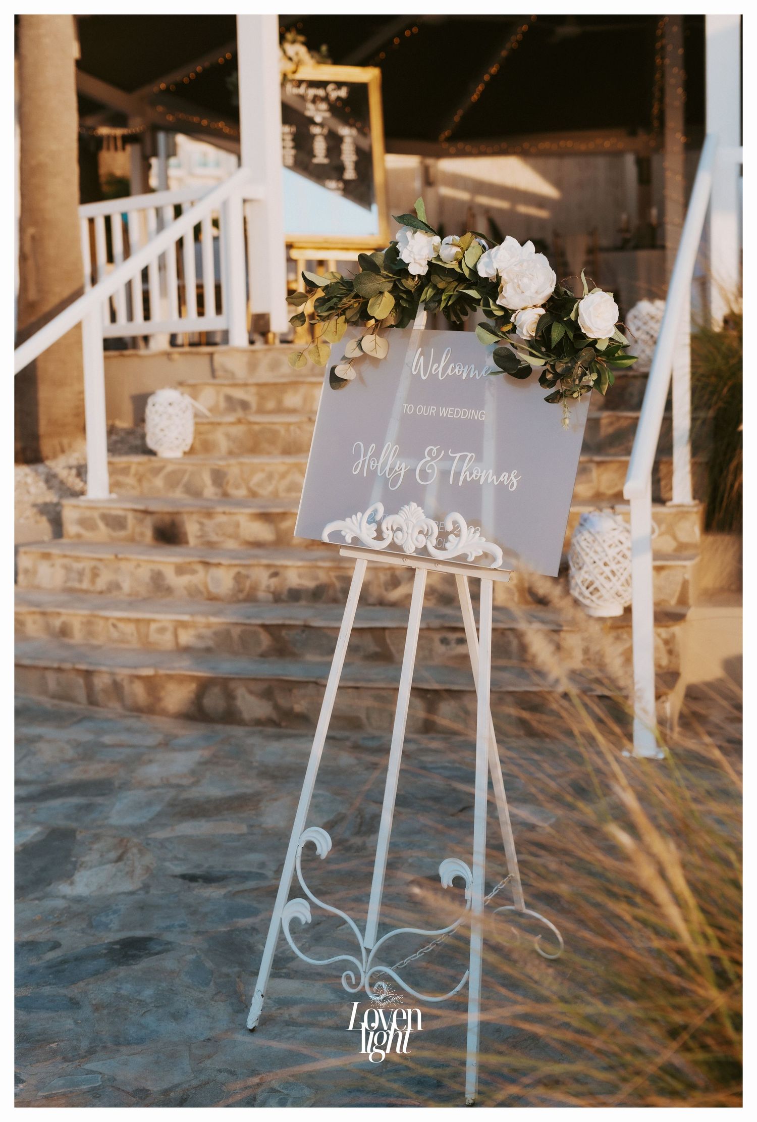 Welcome sign on white easel decorated with white roses and greenery at venue entrance.