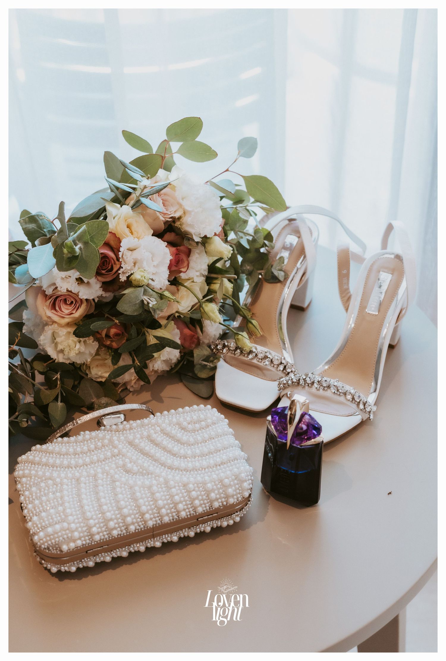 Elegant bridal accessories featuring white heels, beaded clutch, and rose bouquet on table.