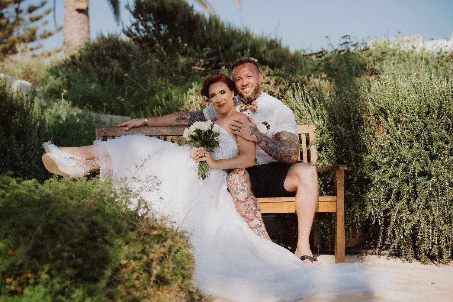 Candid wedding photo of couple relaxing on garden bench amid greenery.