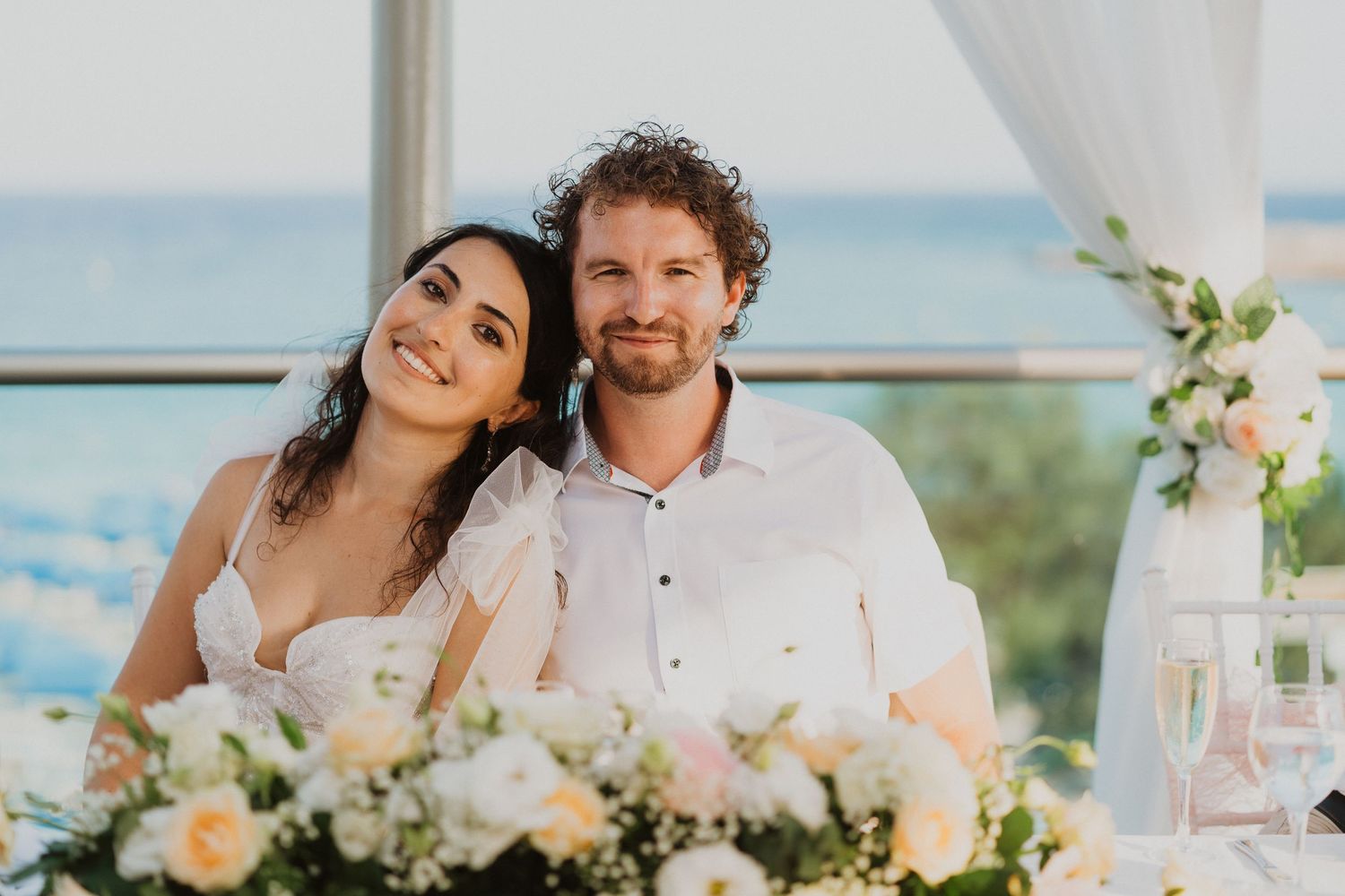 Newlyweds seated at decorated reception table with ocean view and floral arrangements.