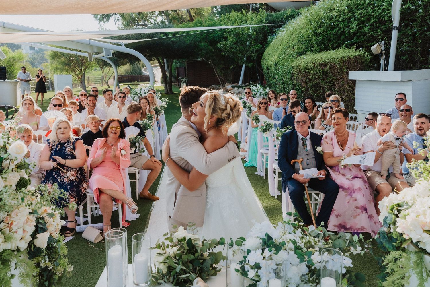 A romantic first kiss at an outdoor wedding ceremony surrounded by joyful guests and floral decorations.
