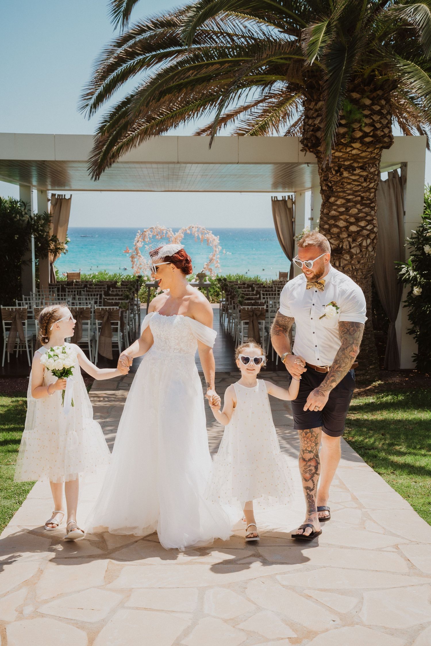 Beachfront wedding ceremony with family walking down palm-lined path under heart-shaped arch decoration.