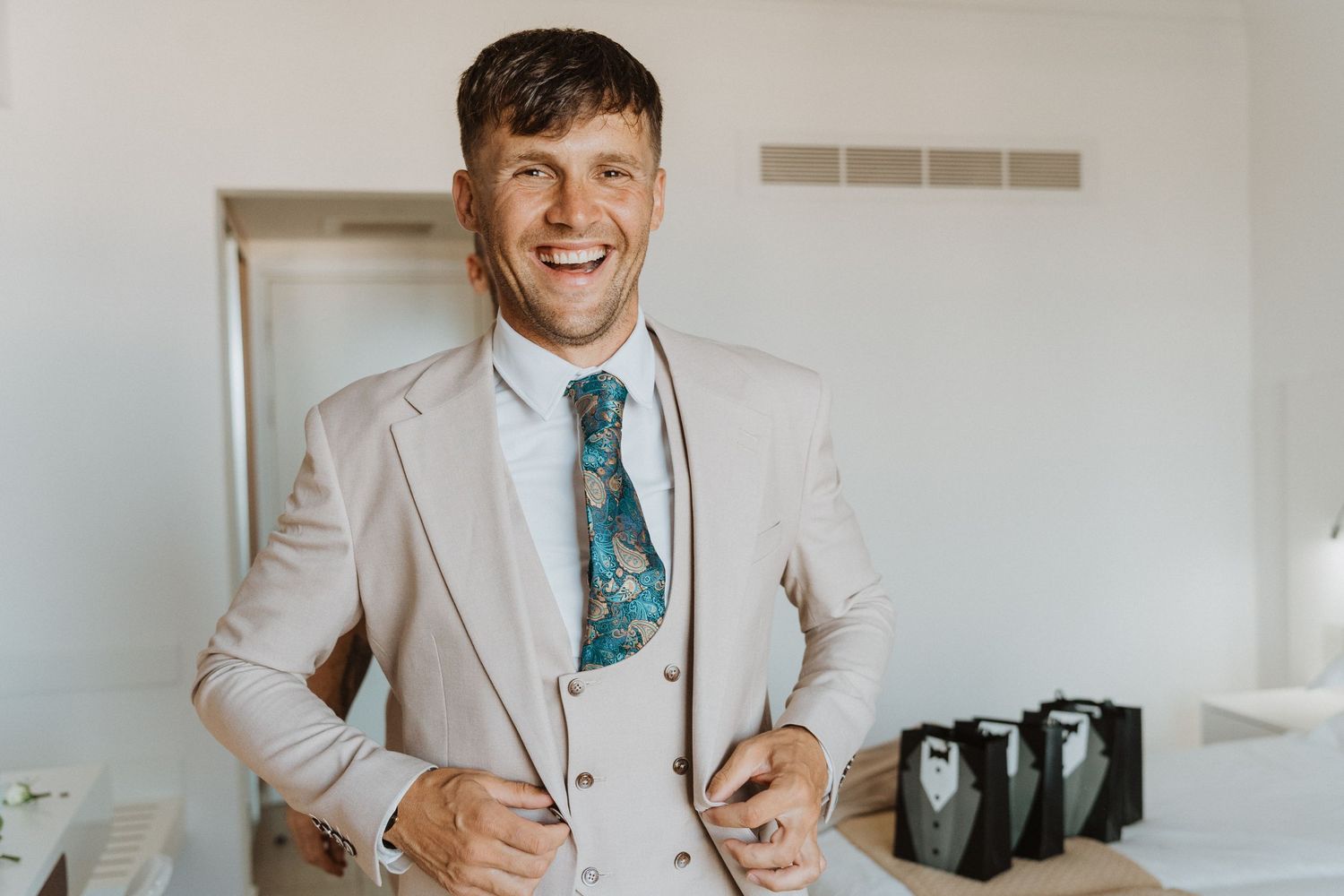 A person wearing a light beige suit and patterned blue tie smiles in a bright white room.