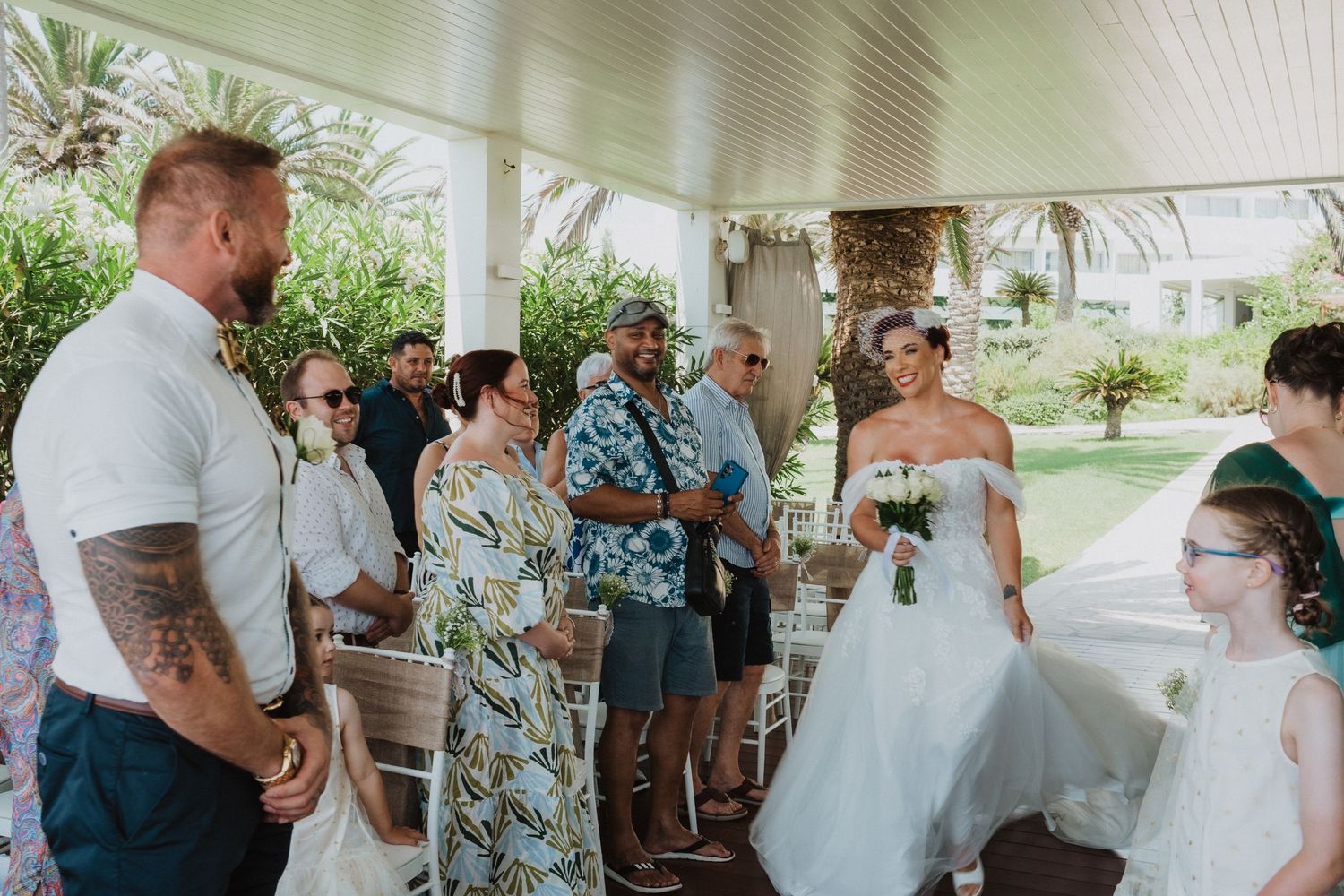 Wedding ceremony attendees gather under a covered outdoor porch in summer attire.