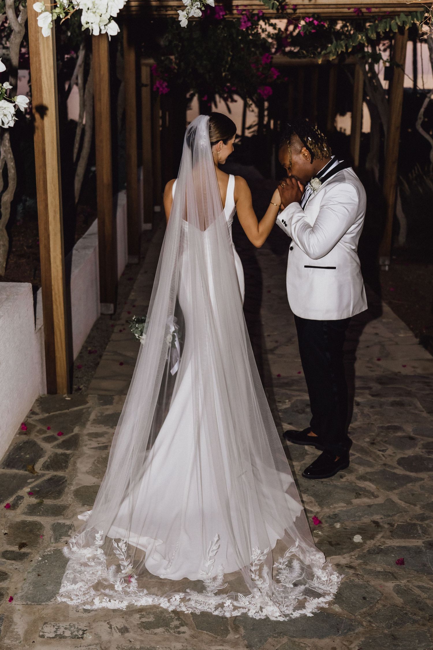 A wedding couple shares an emotional moment under floral decorations in a covered walkway.