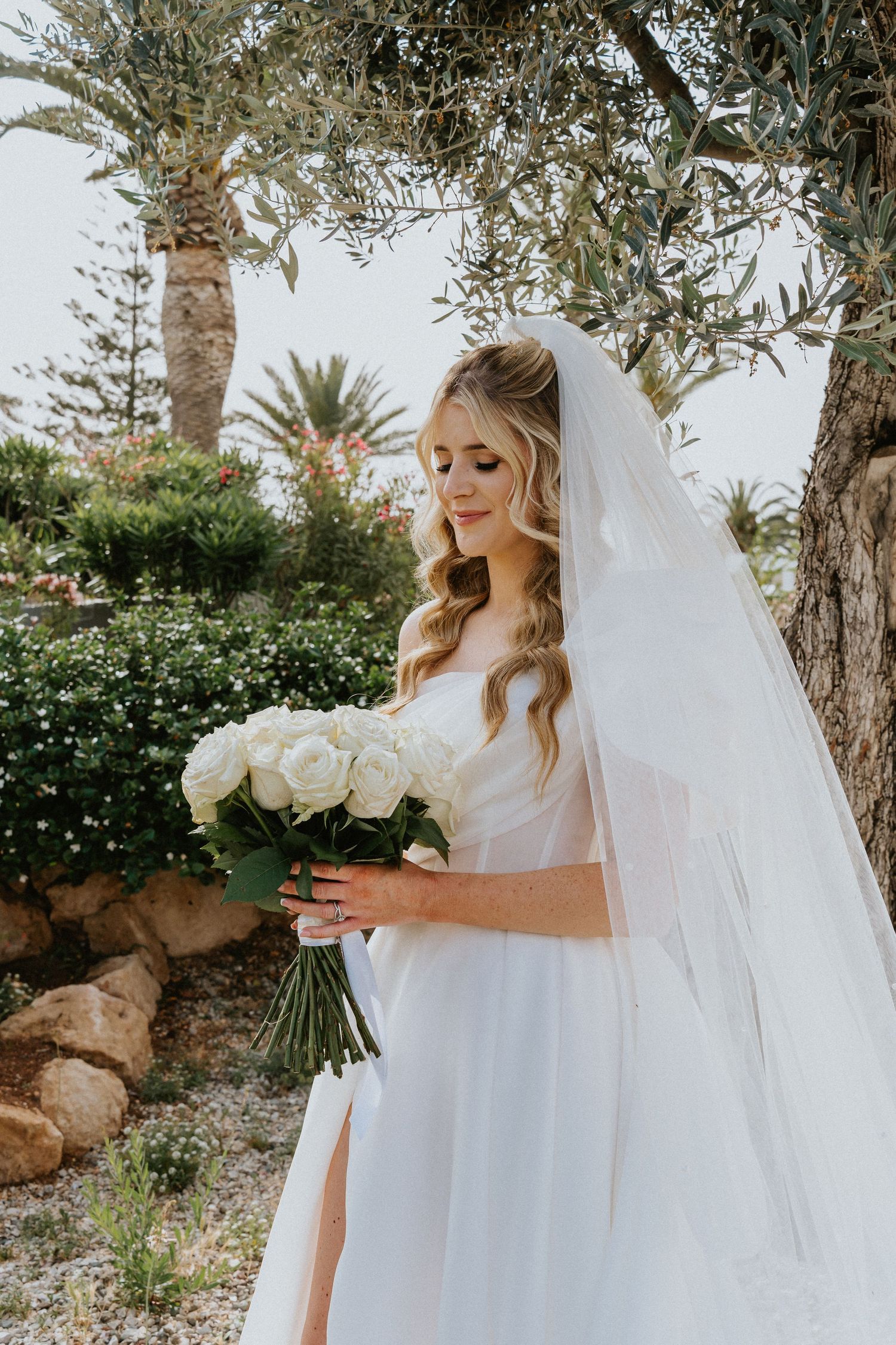 Bride in elegant white wedding dress holding bouquet of white roses stands beneath palm trees and olive branches in garden.
