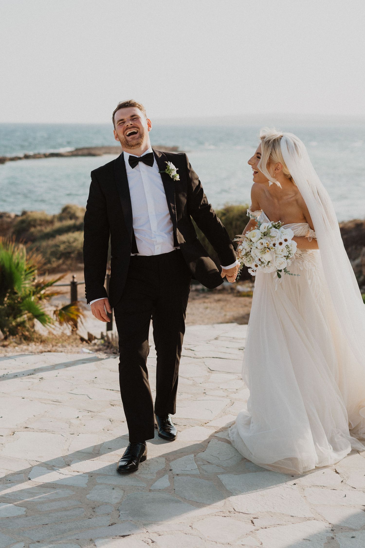 Joyful wedding couple laughs together on seaside stone pathway.