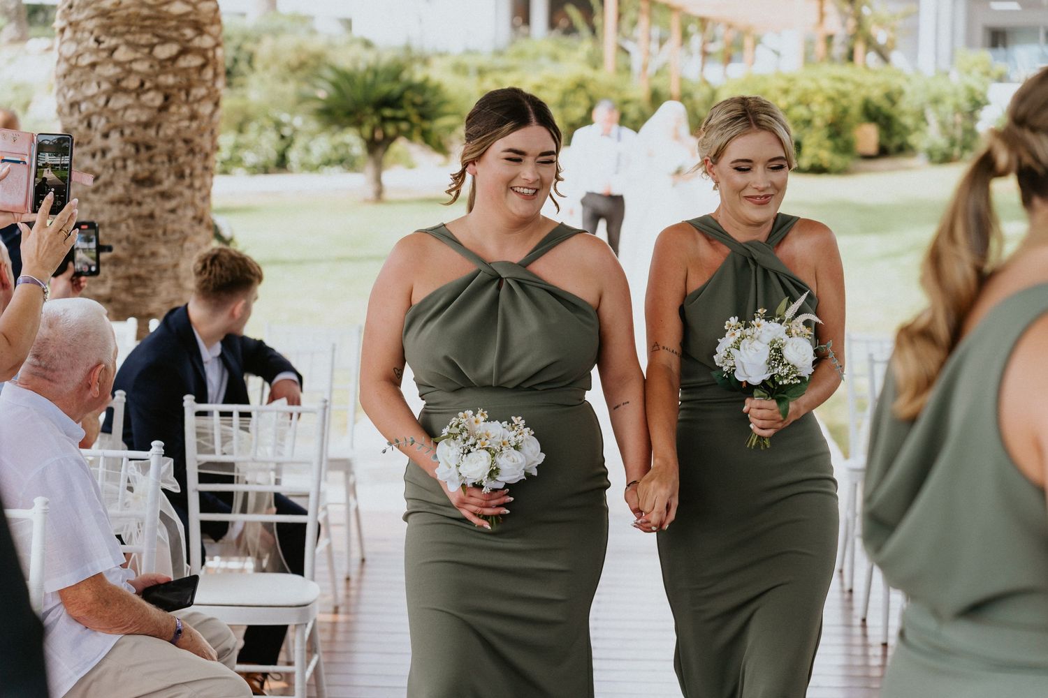 Bridesmaids in sage green dresses proceeding down the aisle with white floral bouquets.