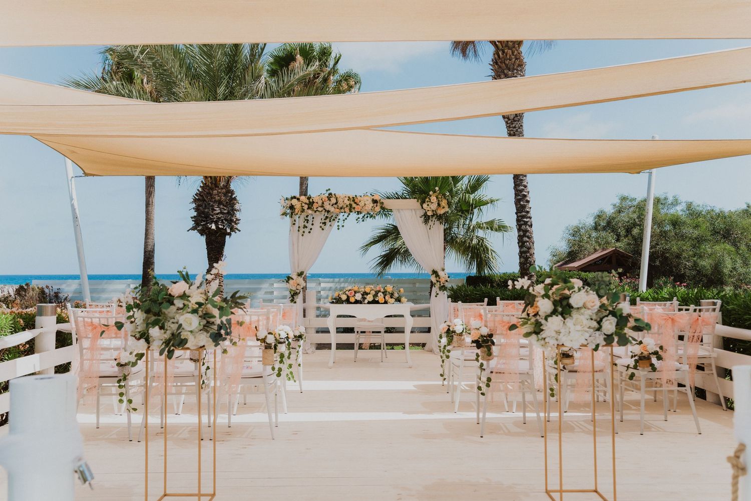 Elegant outdoor beachside wedding ceremony setup with white canopies and floral arrangements on white pedestals.