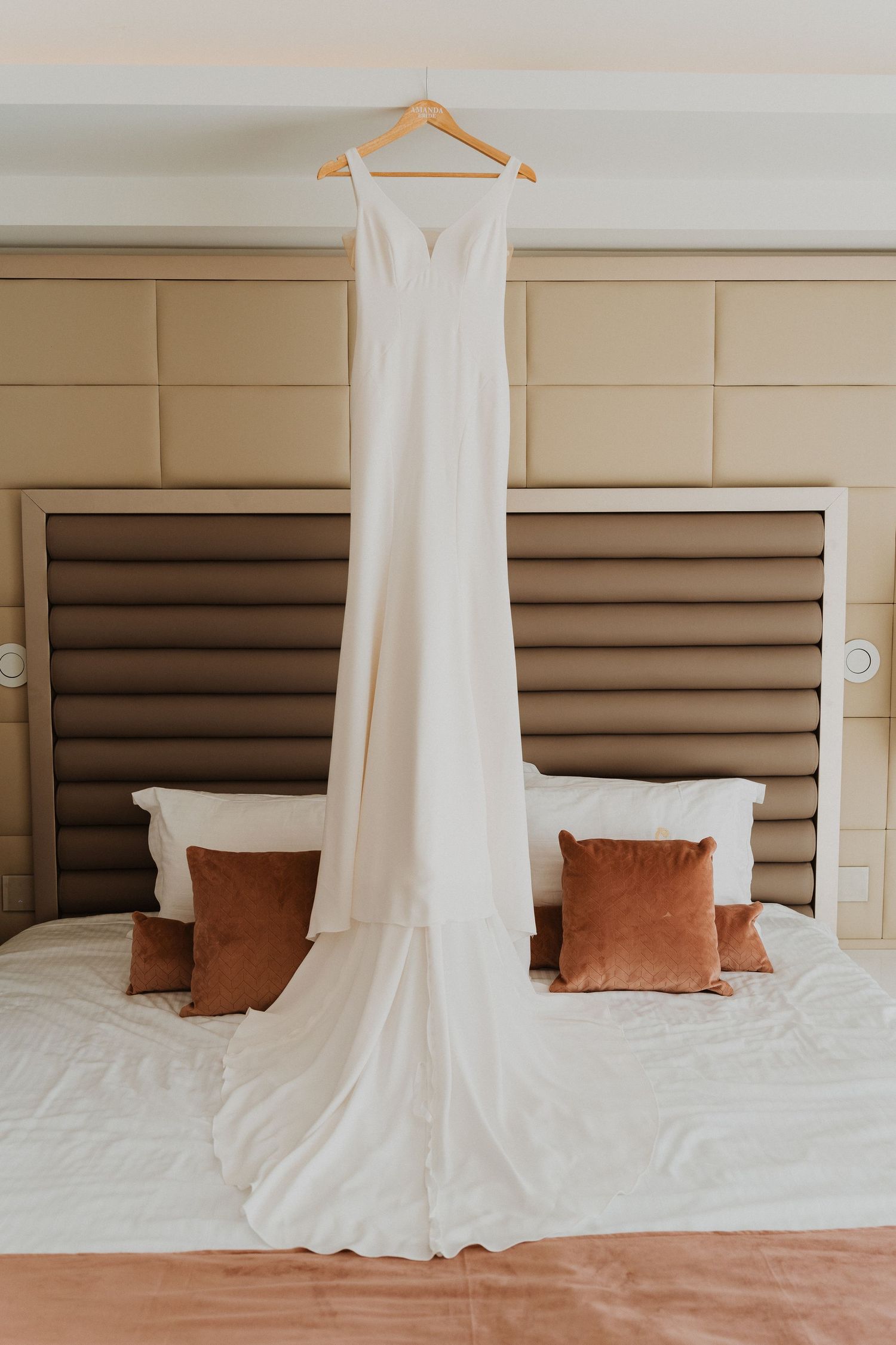 A sleek white wedding gown displayed on a hanger against a neutral bedroom wall.