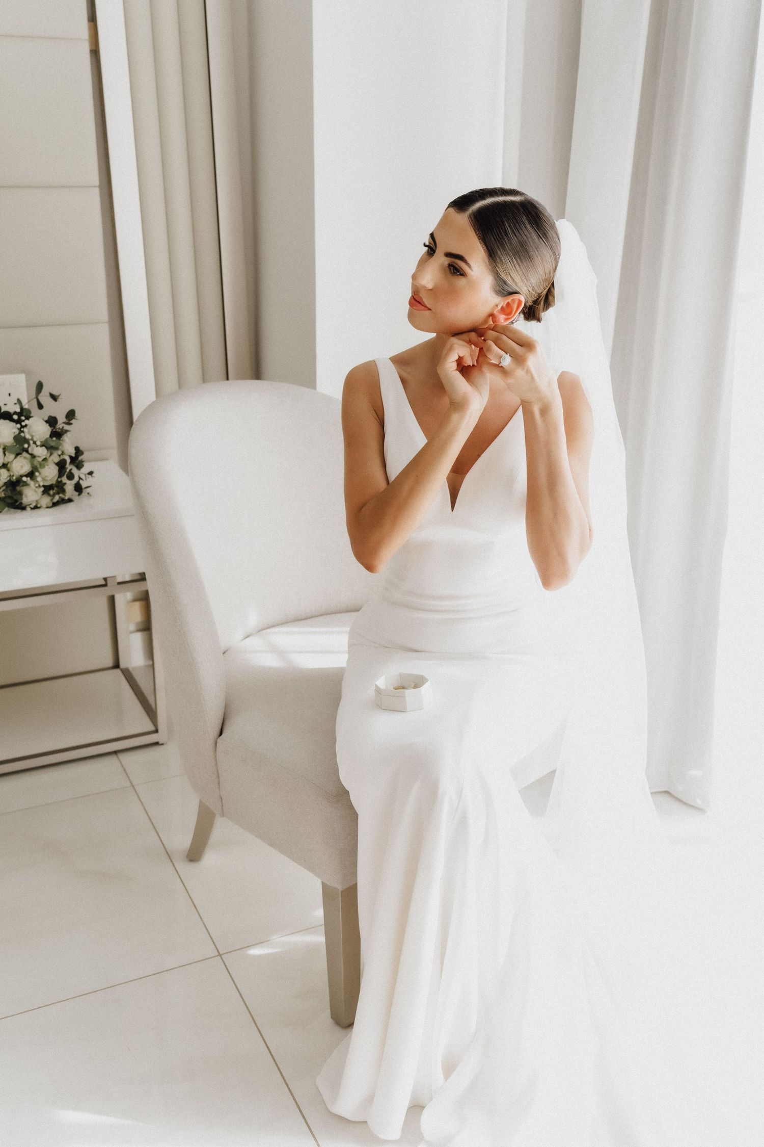 Elegant bride in white dress and veil putting on earrings in a bright room.