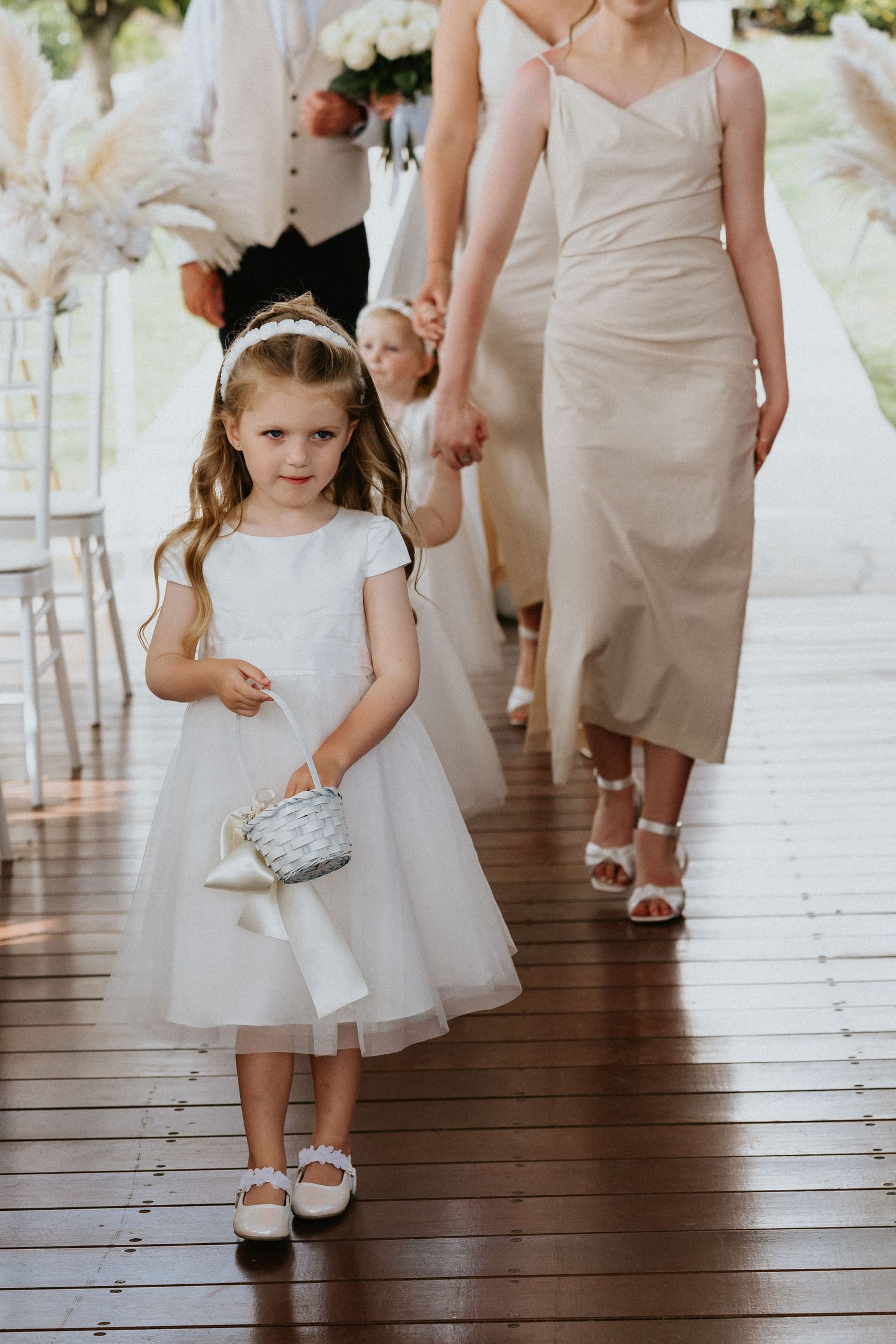 Young flower girl in white dress carries small basket while walking down wooden aisle.