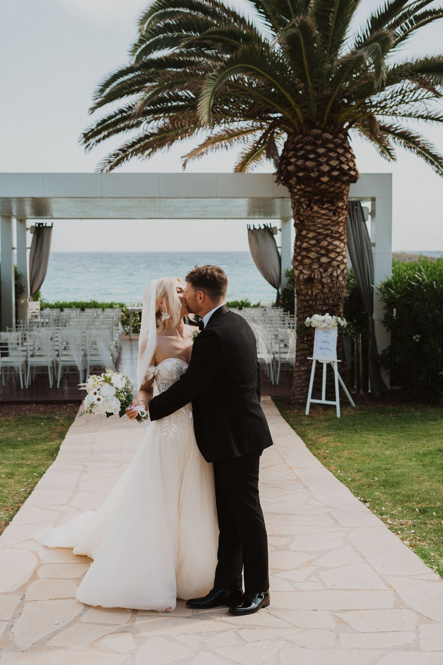 Romantic wedding portrait under a palm tree with ocean views and white ceremony chairs.