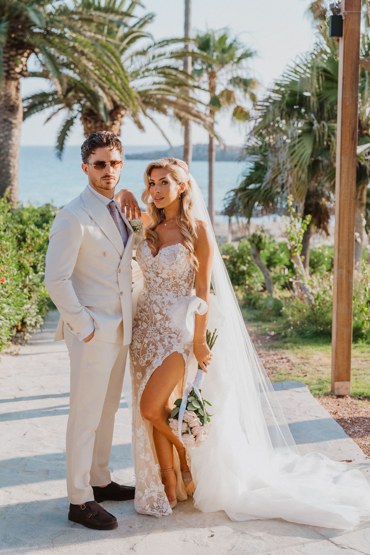 A couple poses in elegant wedding attire with palm trees and ocean views at a tropical destination wedding.