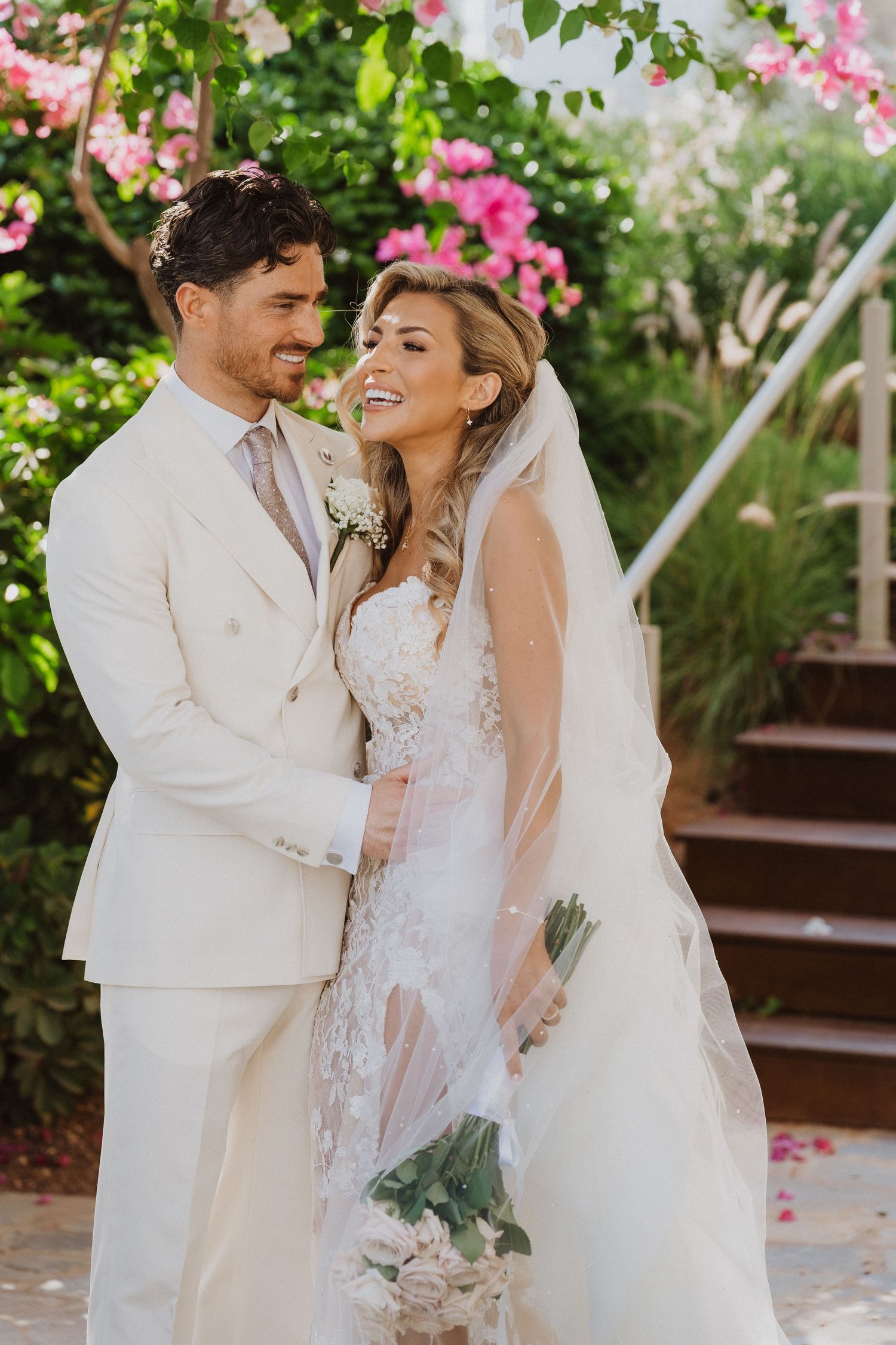 A joyful wedding couple shares a romantic moment in a garden setting with pink bougainvillea flowers and stone steps.