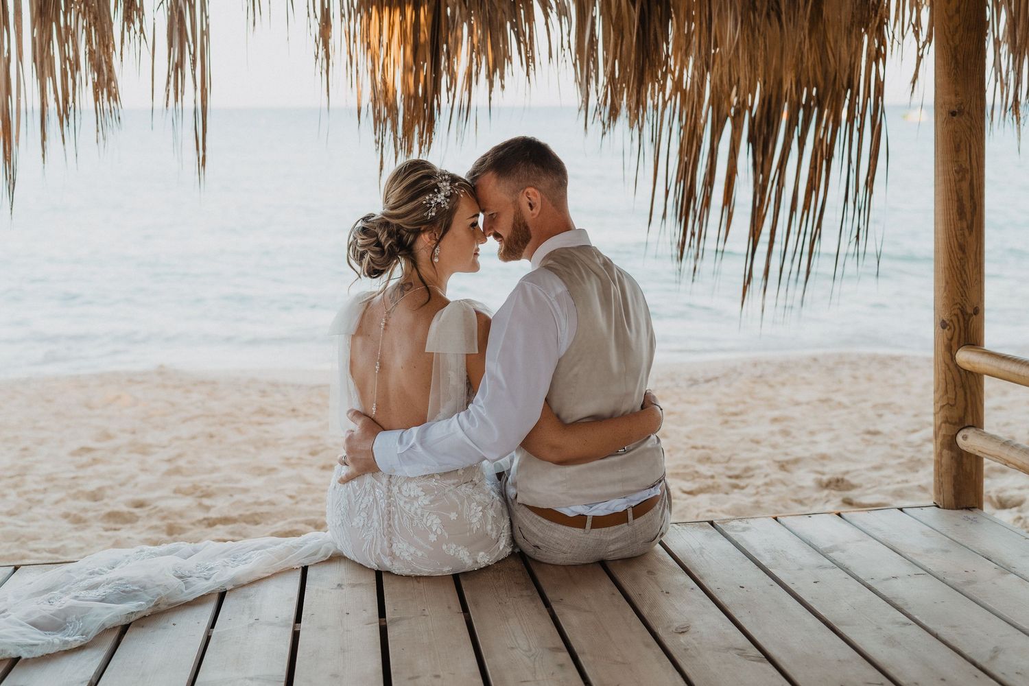Newlyweds share an intimate moment under a thatched palapa on a tropical beach at sunset.