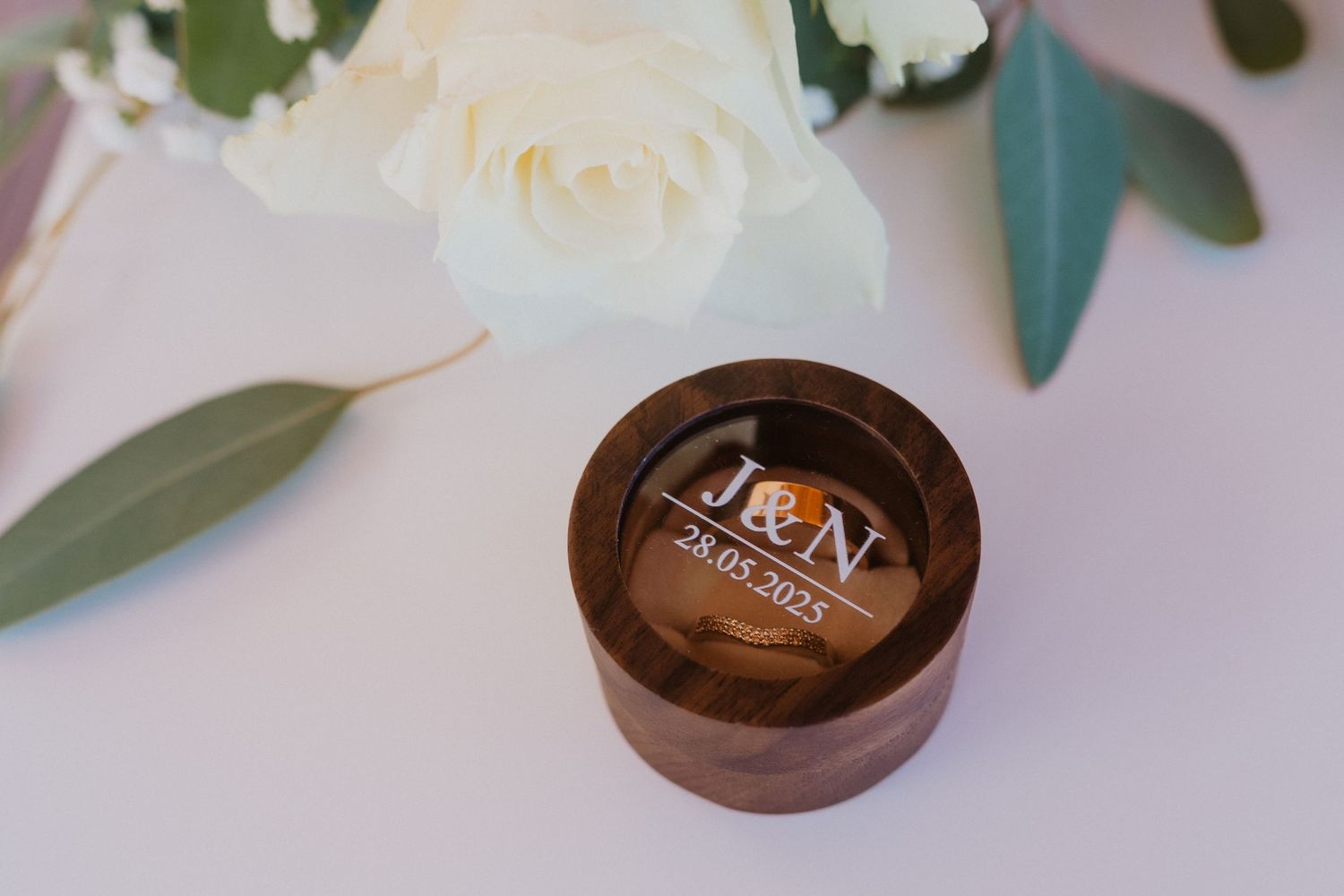 Wooden ring boxes with delicate white roses and eucalyptus leaves arranged on a soft pink background.