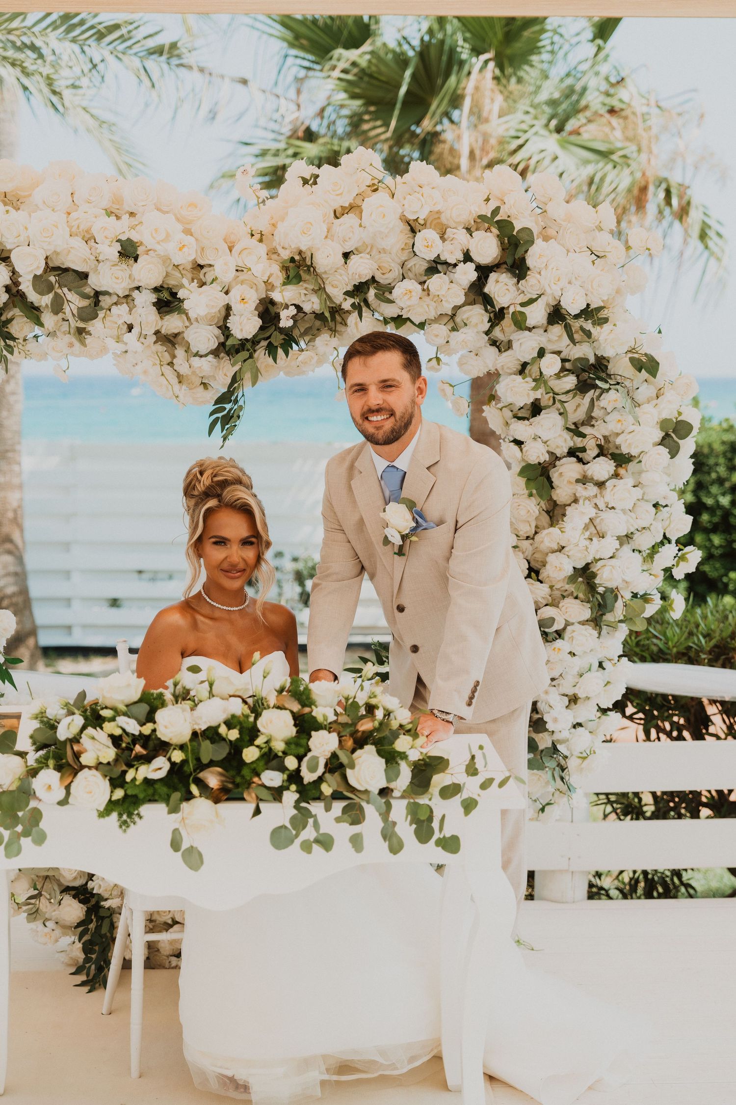 Bride and groom posing under a white floral arch at an outdoor beach wedding ceremony with lush greenery.
