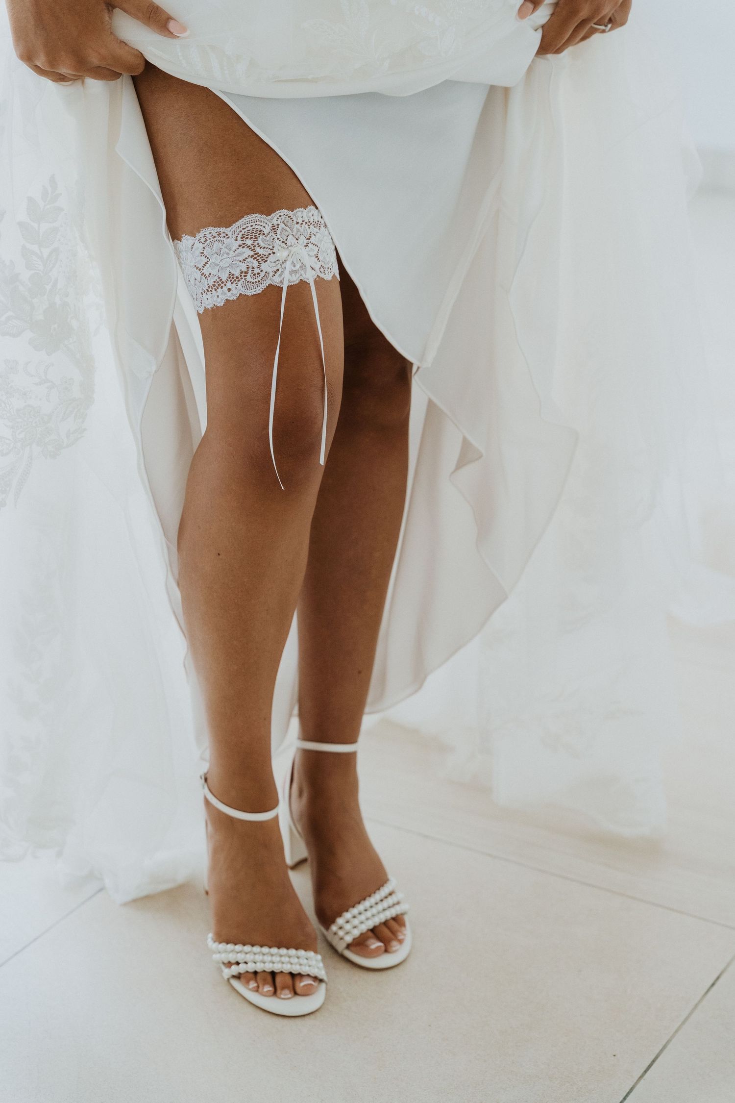 Close-up of bride wearing white lace garter and pearl-embellished sandals with her wedding dress flowing around her legs.